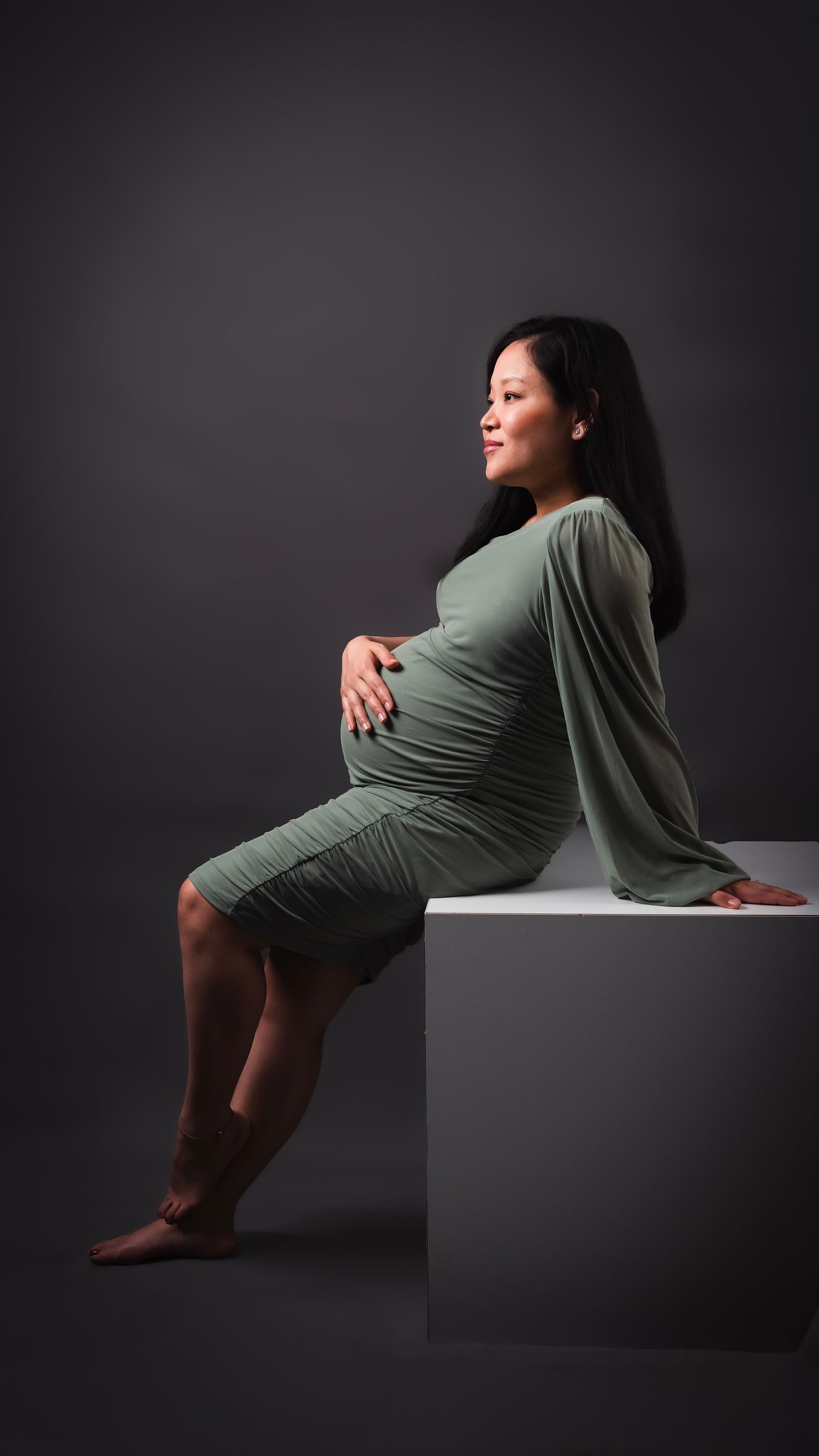 Expectant mother in sage green dress poses elegantly on cube against dark background.