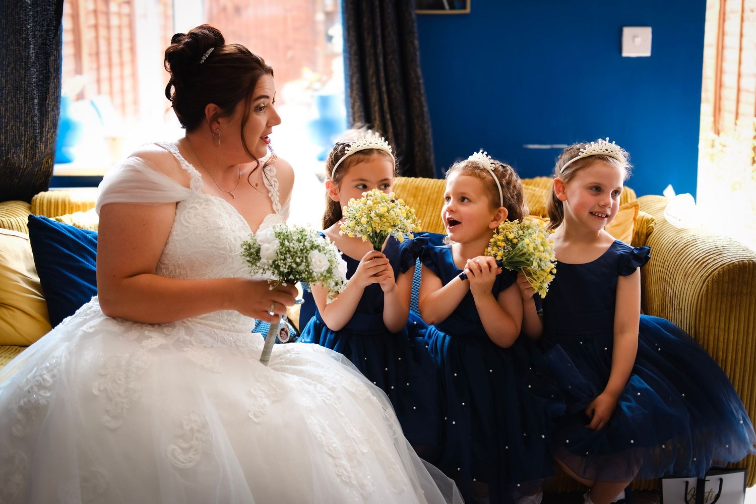 Wedding party shares excited moment on blue couch with yellow bouquets.