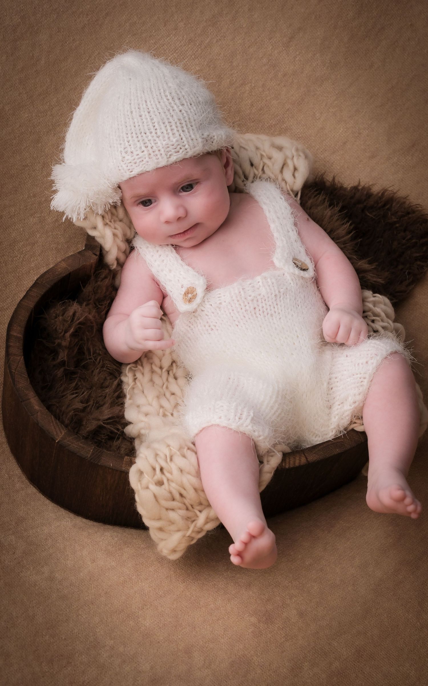 Baby wearing white knit outfit resting in wooden bowl with cream colored textures.