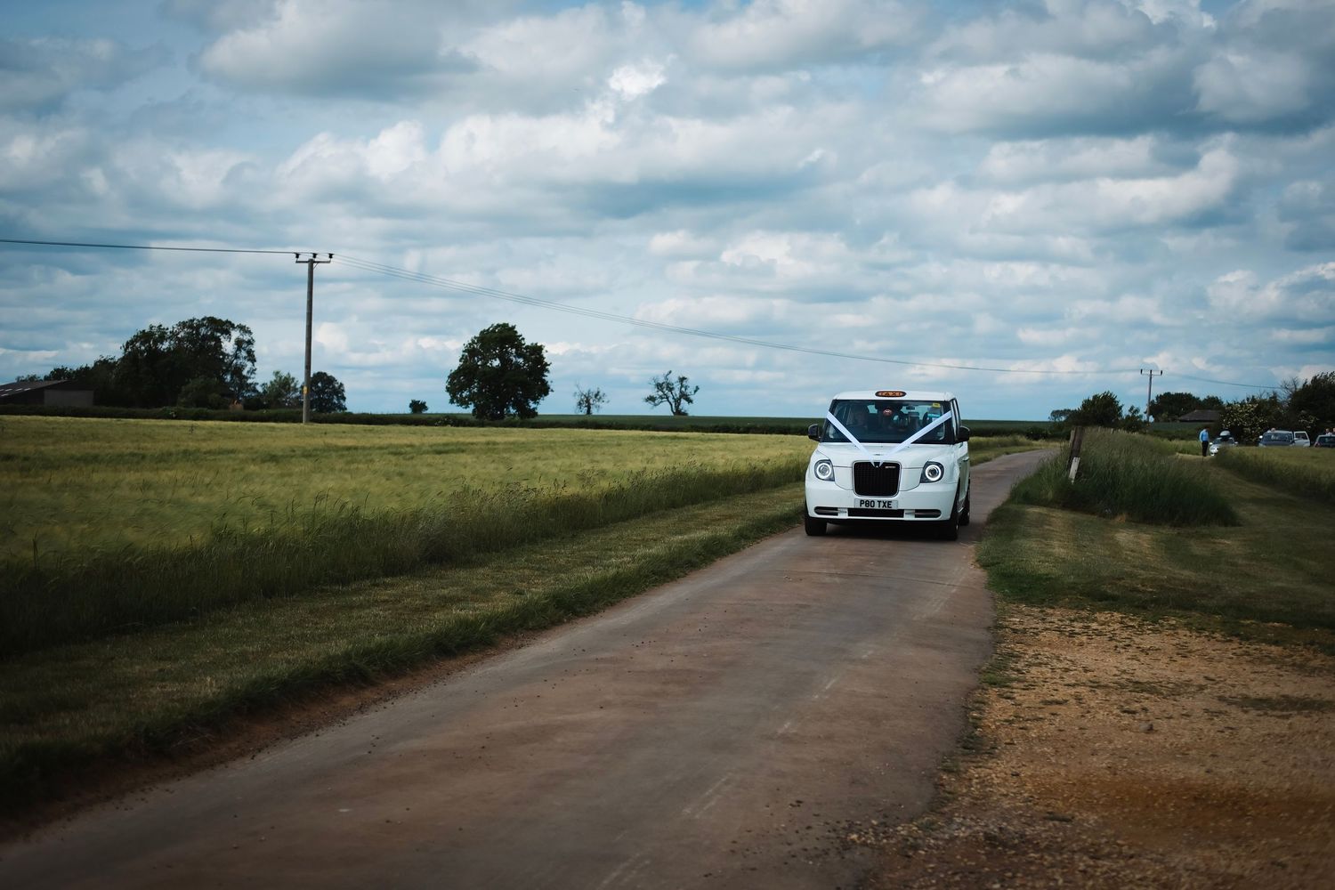 Rural country road stretches into distance under dramatic cloudy sky with cars visible in background.