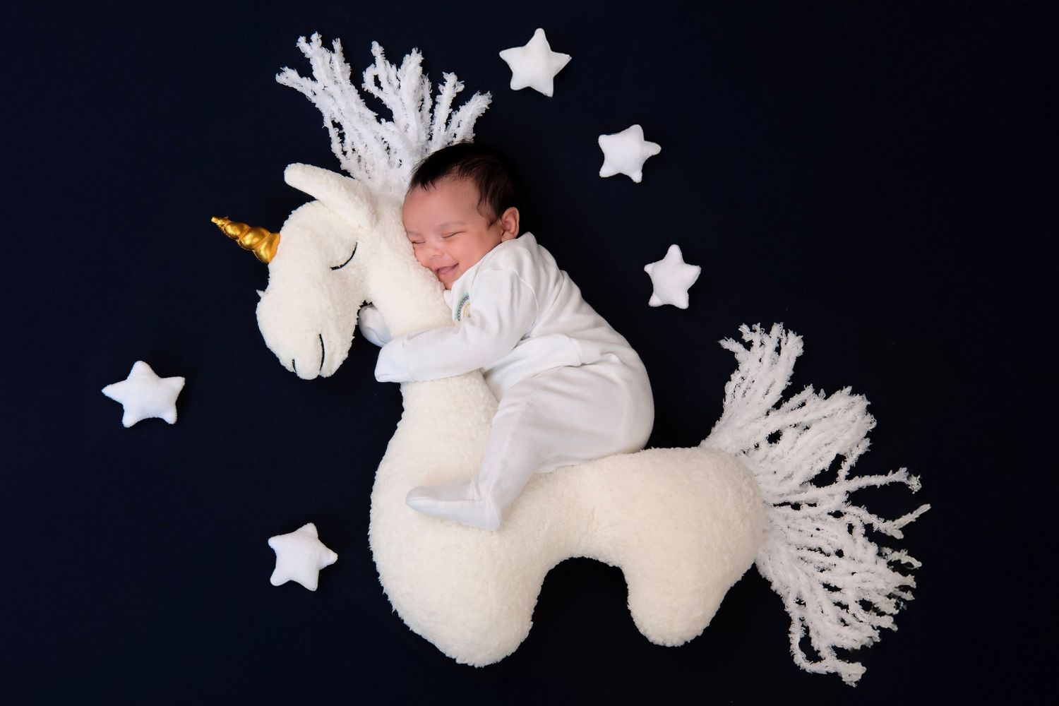 Sleeping newborn posed with white plush unicorn toy and star decorations against black background.