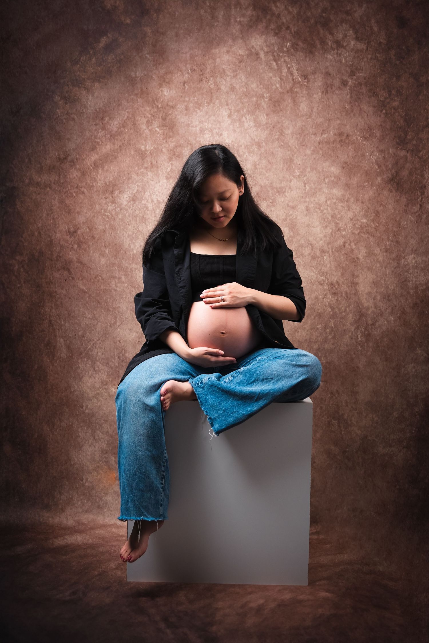 Maternity portrait with mother wearing black top and denim against textured brown backdrop.