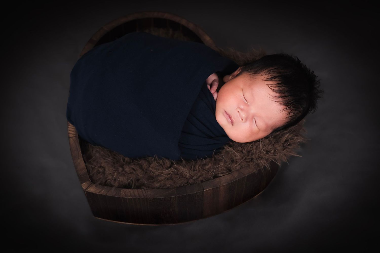 Newborn baby sleeping peacefully in a dark wooden box with soft brown fabric.