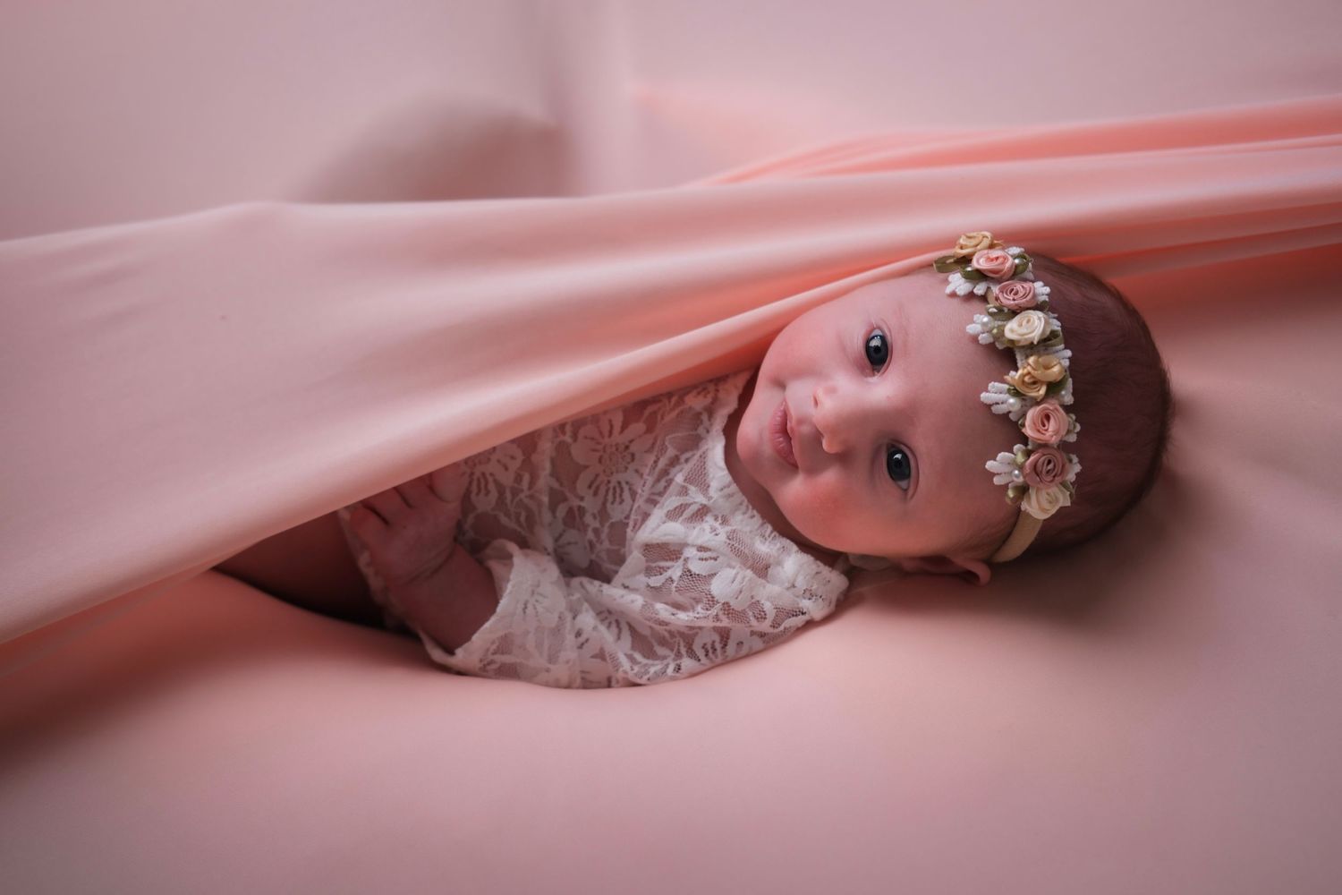 Baby with floral headpiece nestled in flowing pink fabric during photo session.