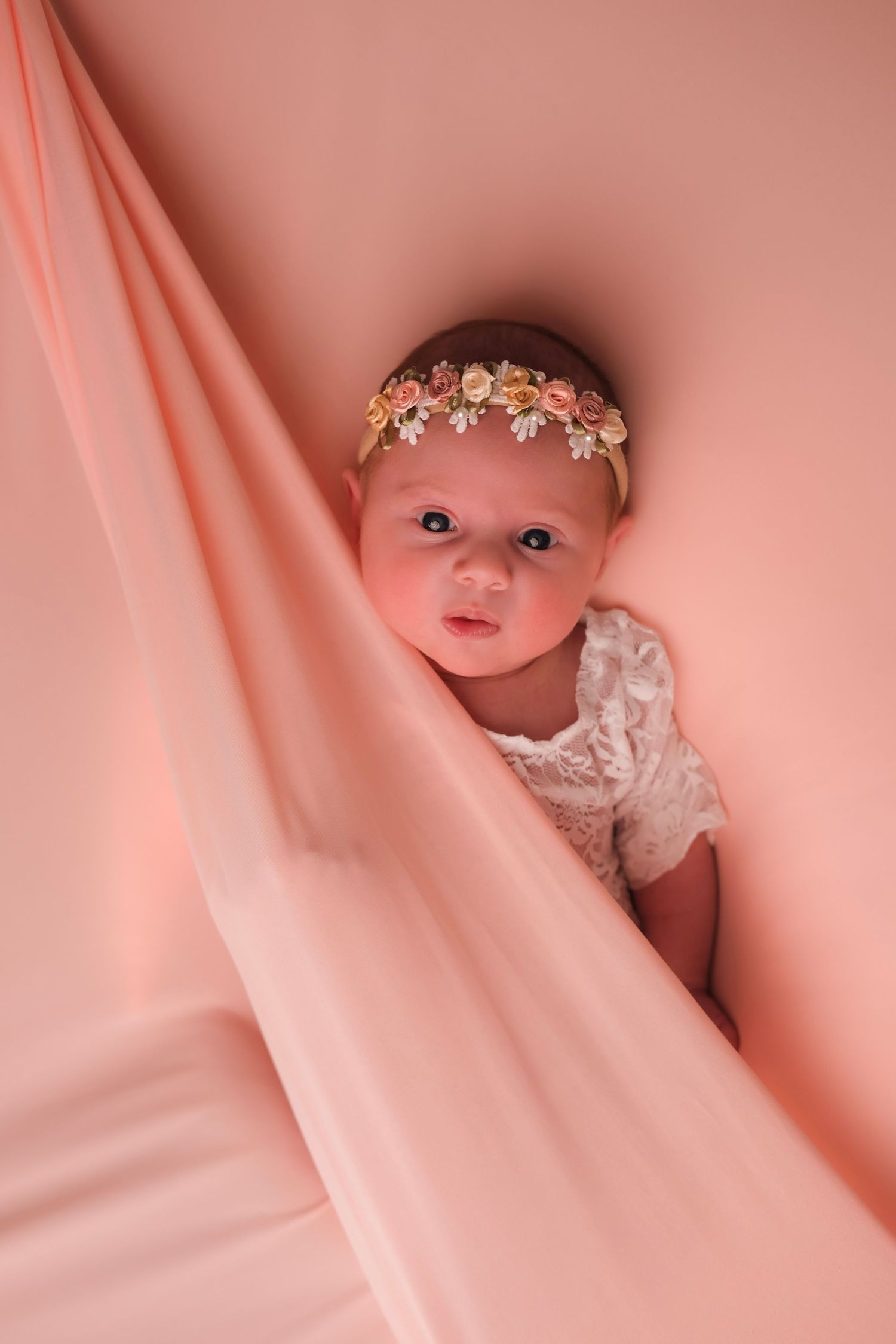 Baby wearing floral crown peeks through pink fabric in dreamy portrait.