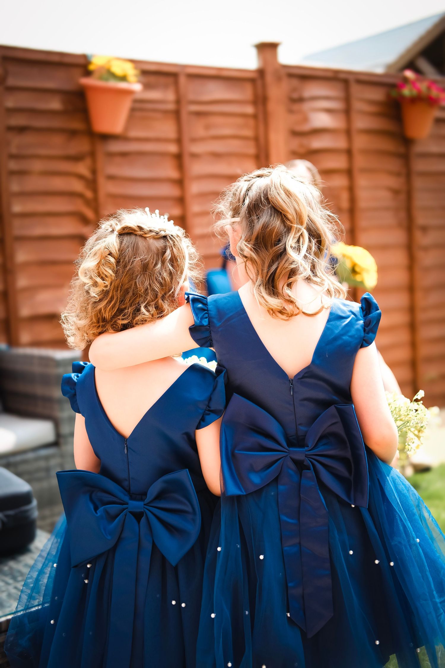 Two matching navy blue dresses with large bows and tulle skirts shown from behind against a wooden fence.