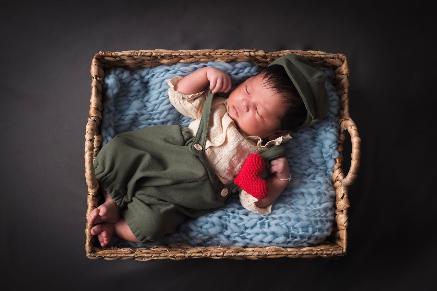 Baby rests in rustic woven basket with soft blankets and plush toy on dark background.