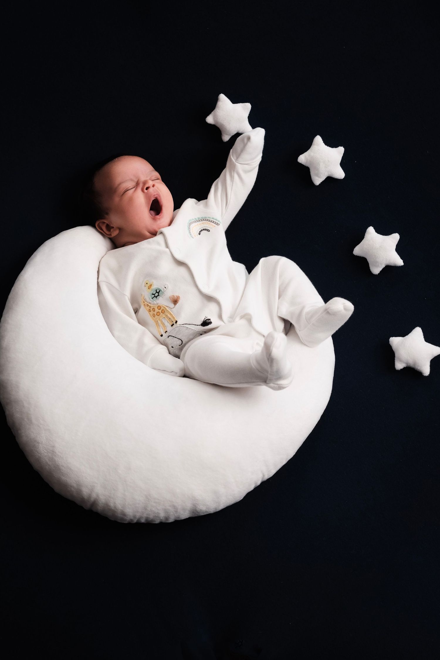 Newborn sleeping on white crescent moon prop with stars against dark background.