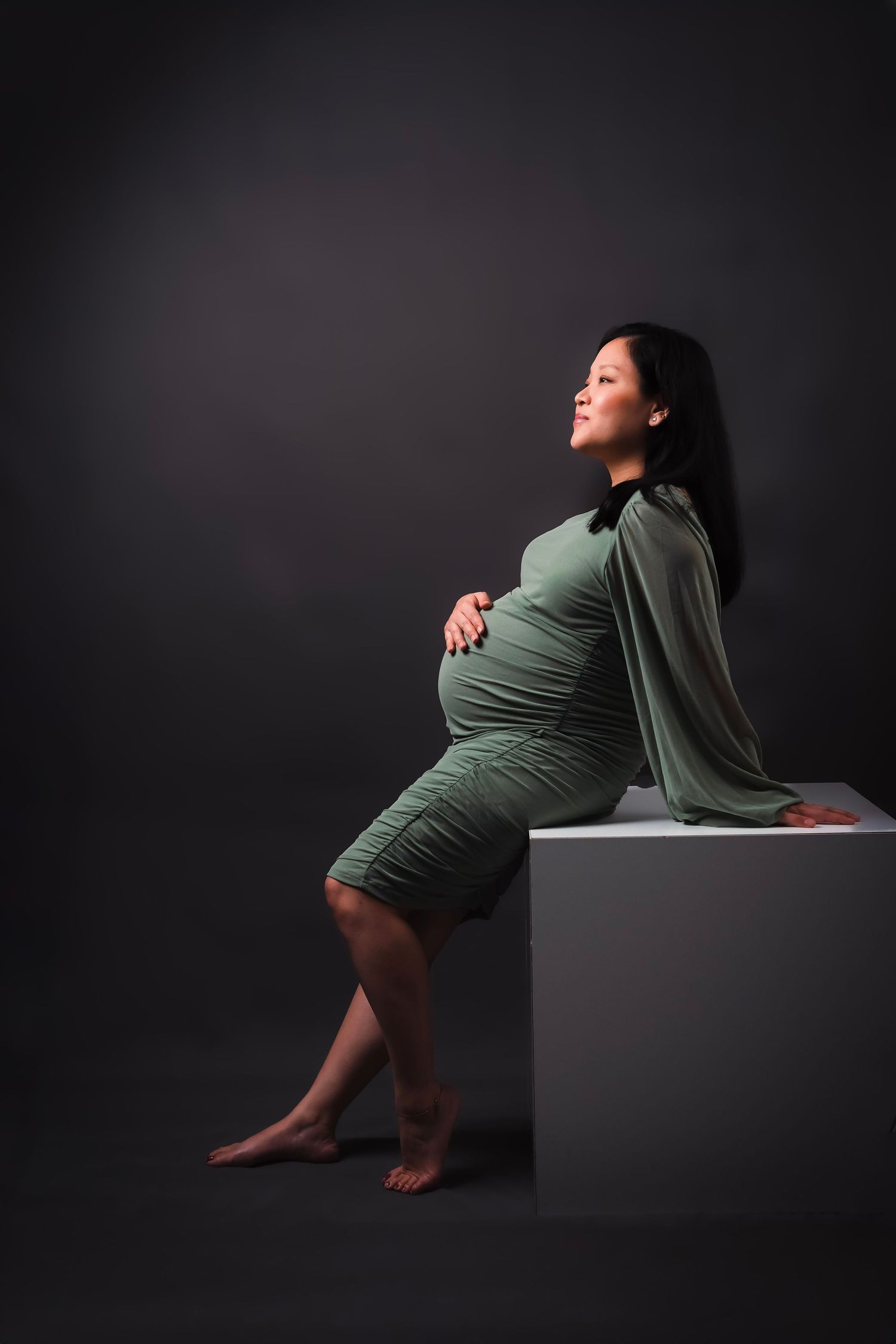 Maternity photoshoot featuring woman in olive dress posing on dark cube against black background.