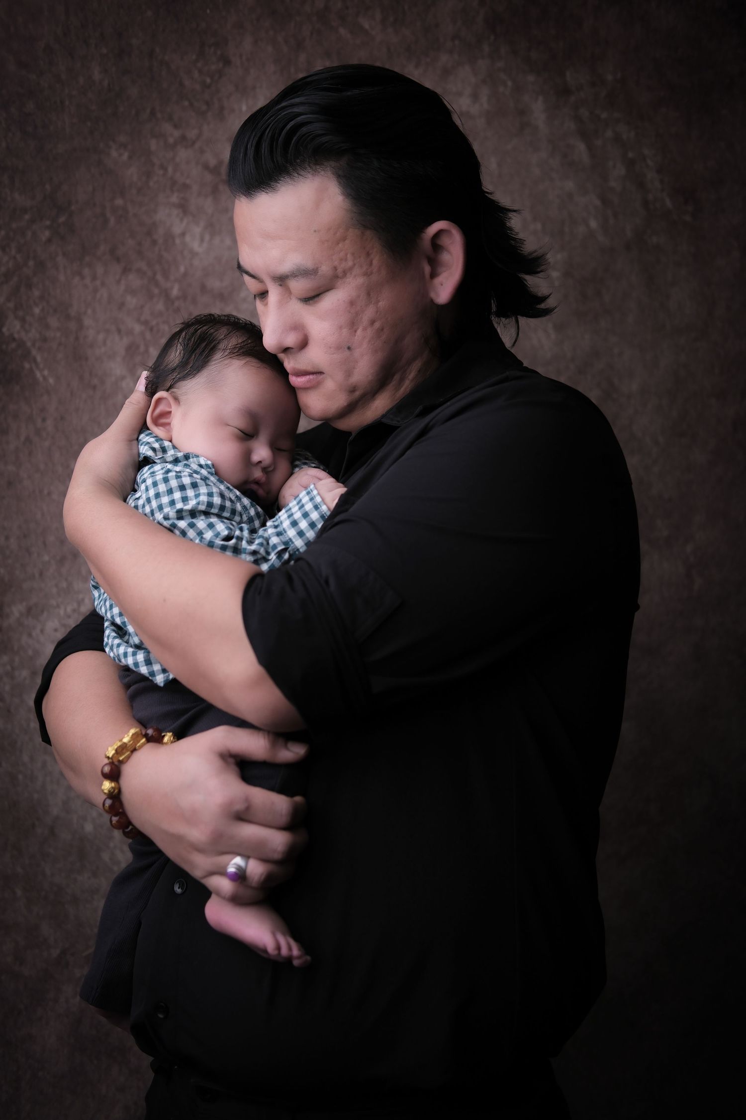 Tender moment captured in dark tones as parent cradles sleeping infant wearing checkered outfit.