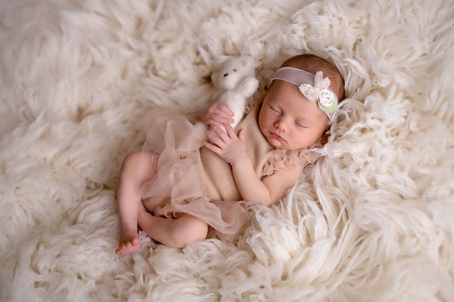 Sleeping newborn in blush tulle outfit cuddles a white stuffed bear on a cream fluffy fur backdrop.