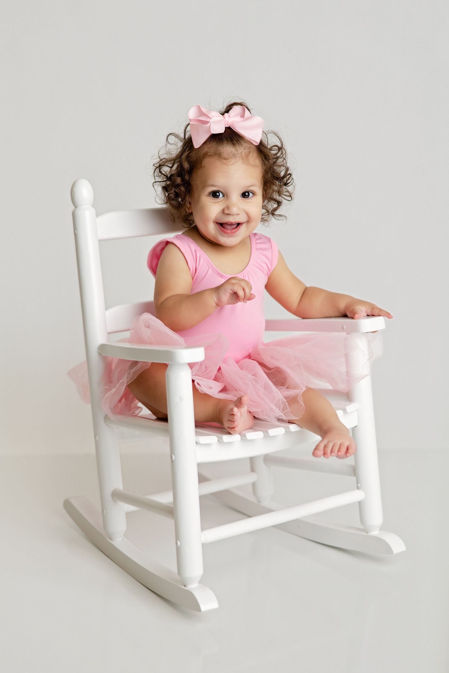 Happy toddler girl in pink tutu and bow sitting in a white rocking chair against grey background.