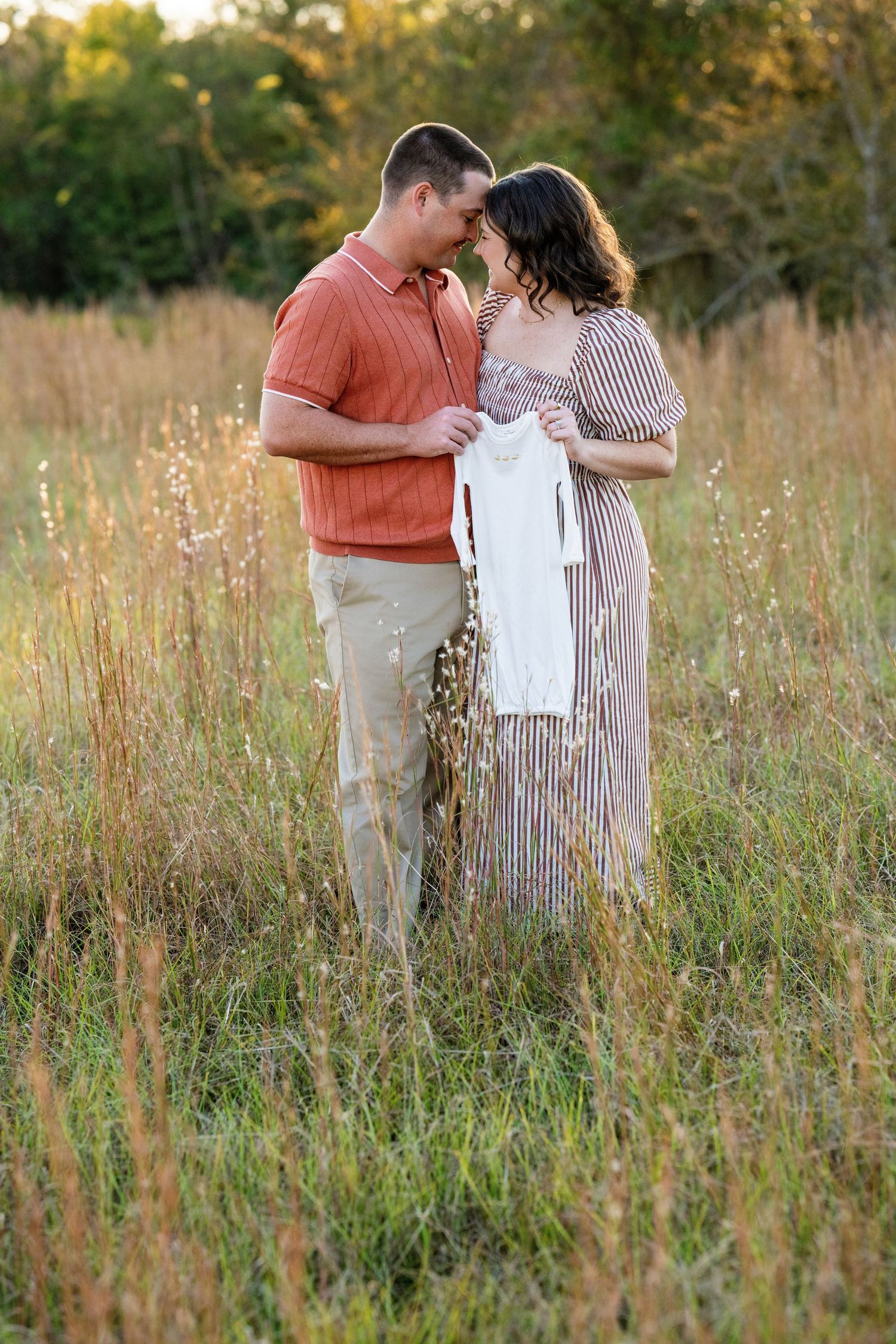 Expecting couple touches foreheads tenderly while holding white baby onesie in sunlit meadow.