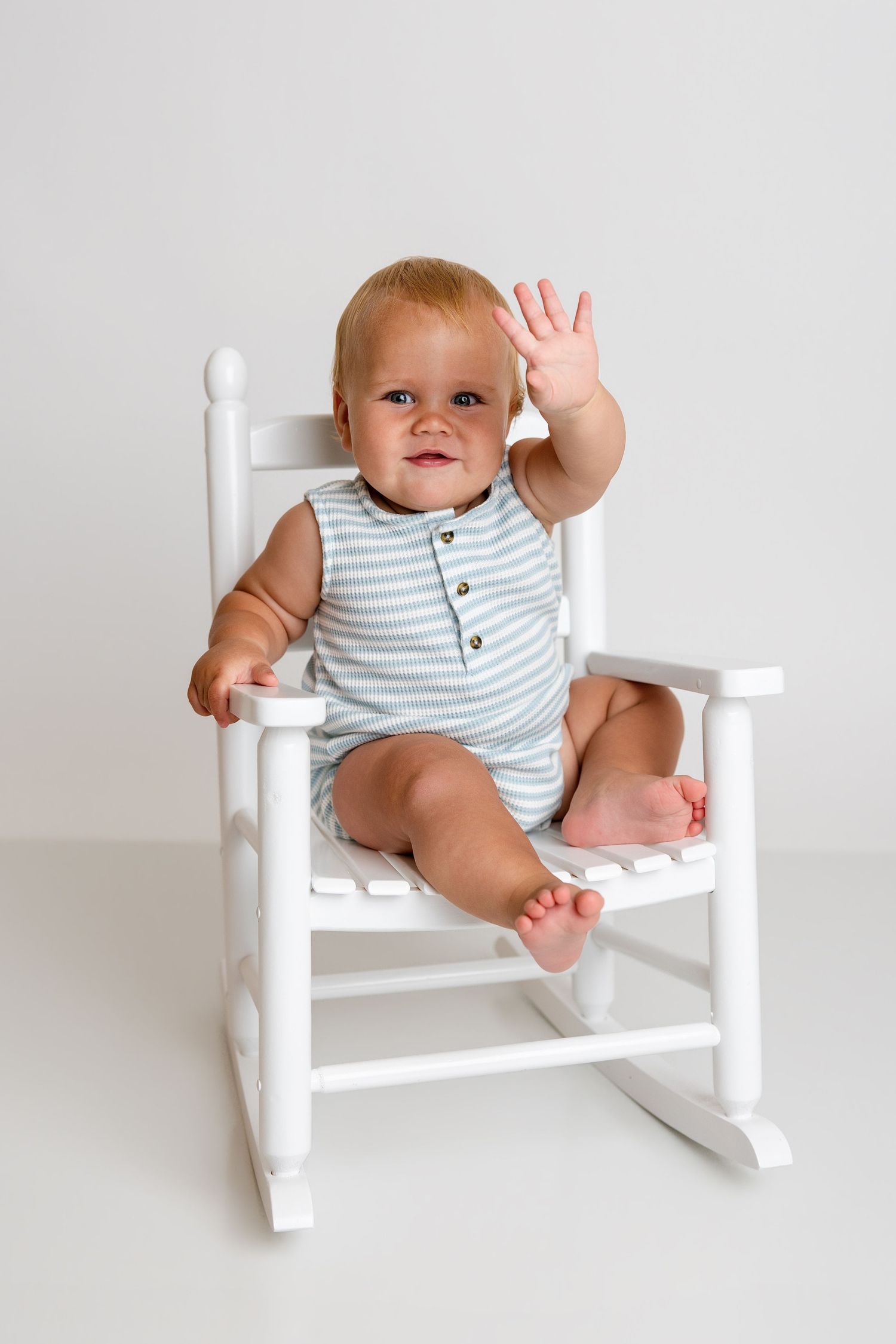 An adorable baby in a striped romper waves while sitting in a small white rocking chair.