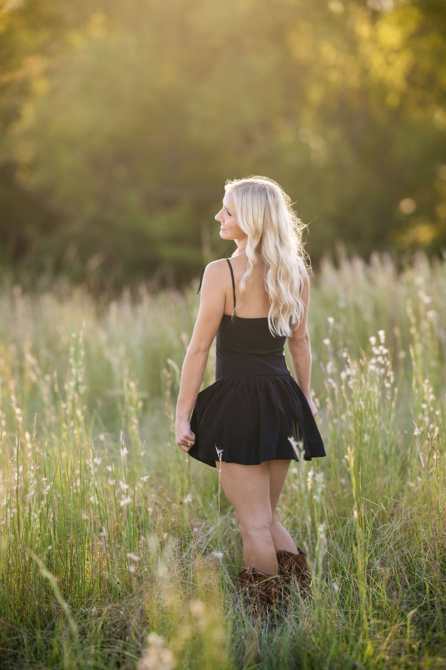 Blonde teen in black dress and brown boots stands in sunlit meadow, back to camera.