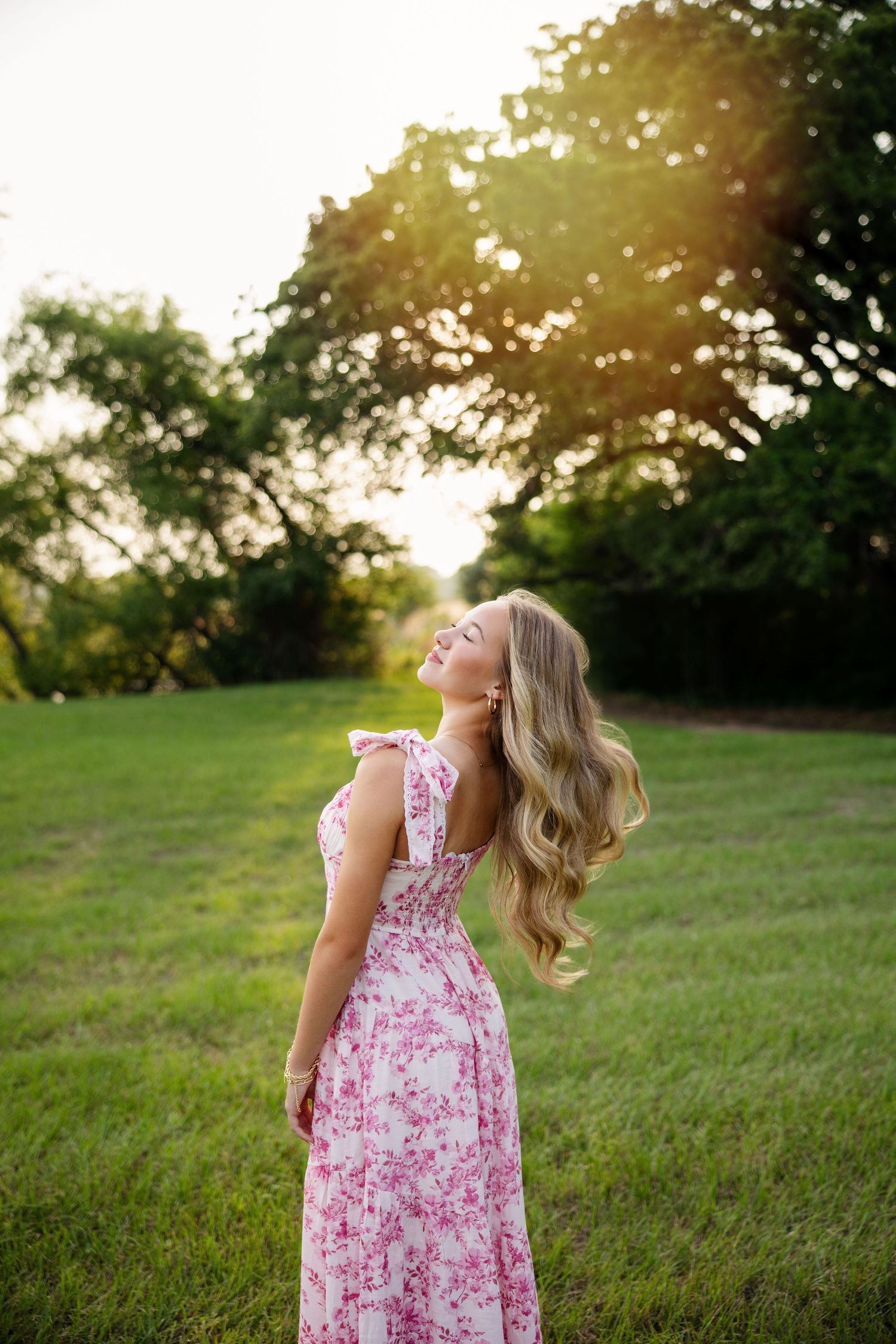 Blonde woman in pink floral maxi dress tosses hair back in sunlit green outdoor setting.