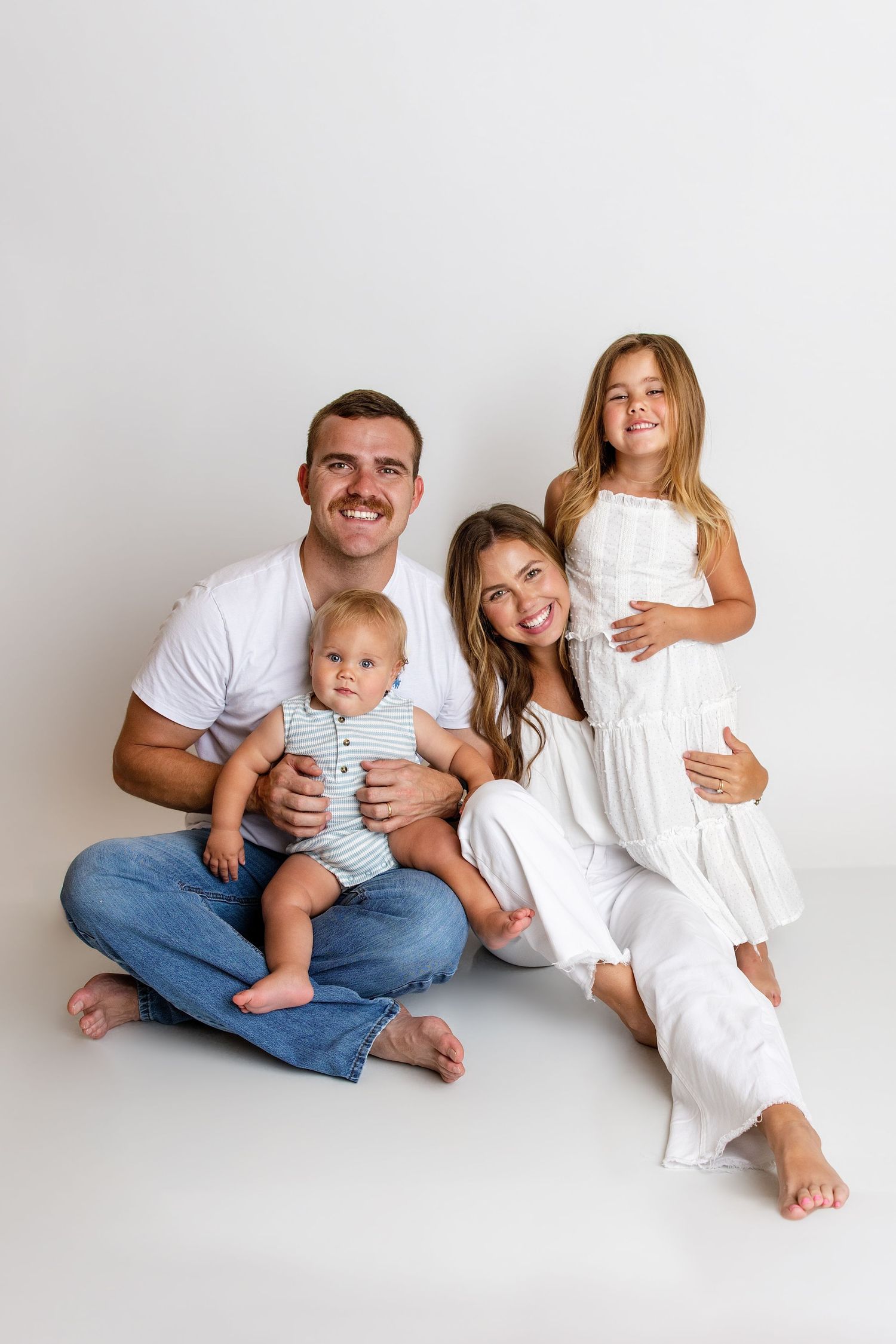 A happy family of four with two young children poses together on a white studio floor.