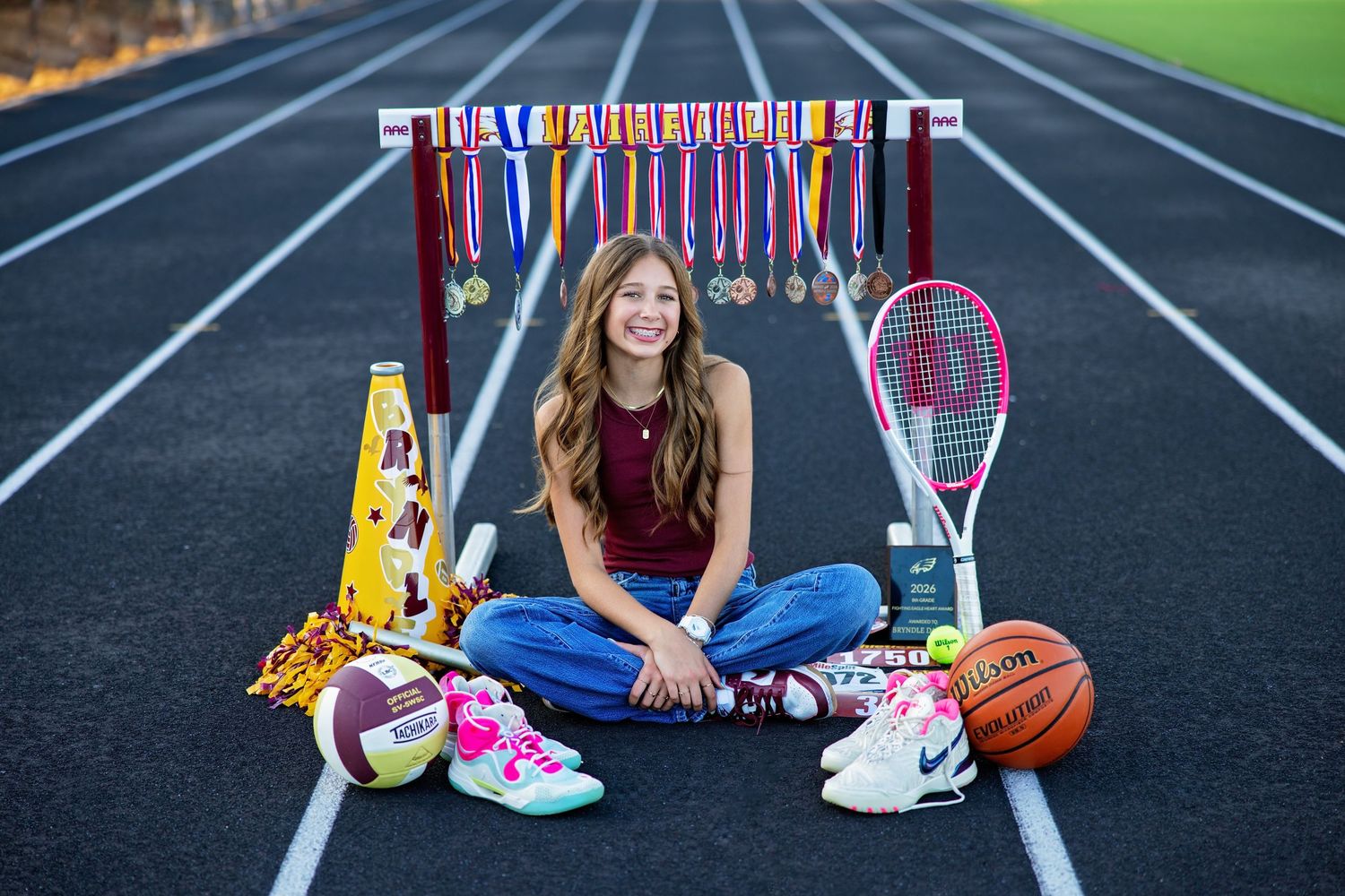 Teen girl sitting on track surrounded by sports equipment, medals, and athletic gear.