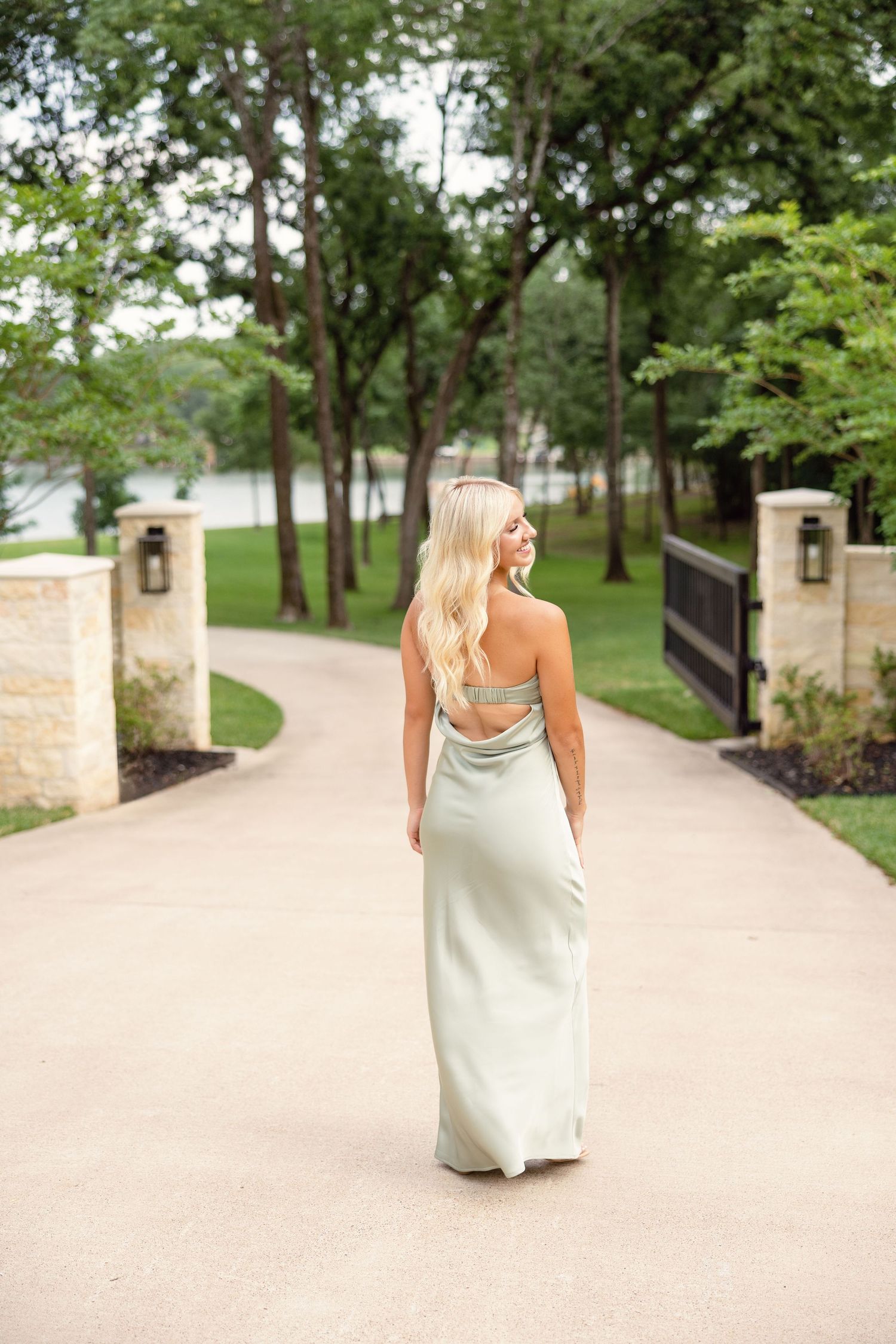 Blonde woman in sage green halter gown walks along elegant tree-lined driveway.