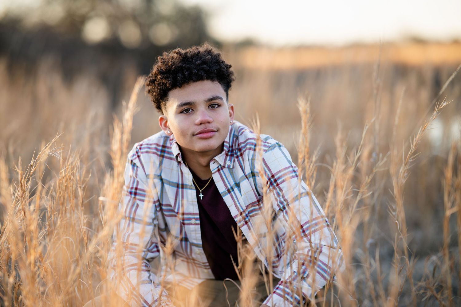 Curly-haired teen boy in plaid shirt poses in golden dry grass field for senior photos.