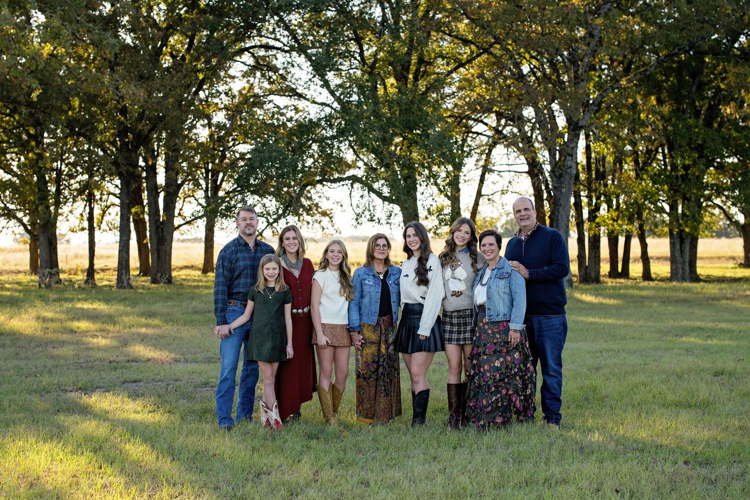 Large family group of nine smiling together in a sunlit green field with trees.