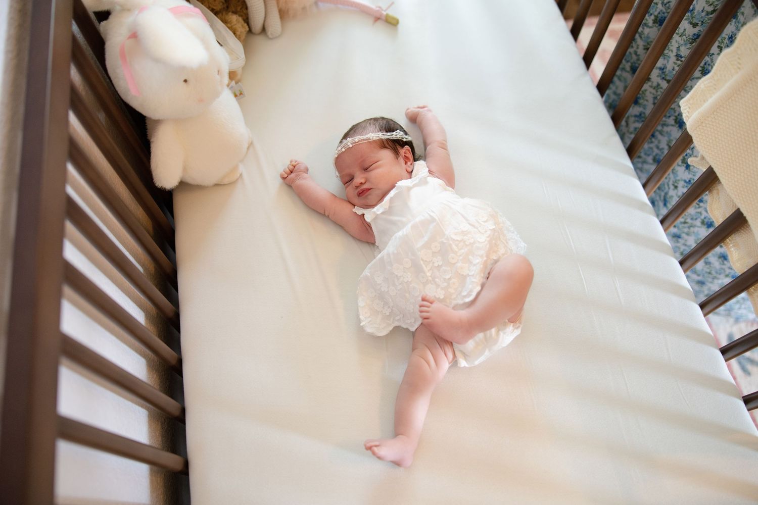 Newborn baby girl in white lace outfit sleeping peacefully in wooden crib.