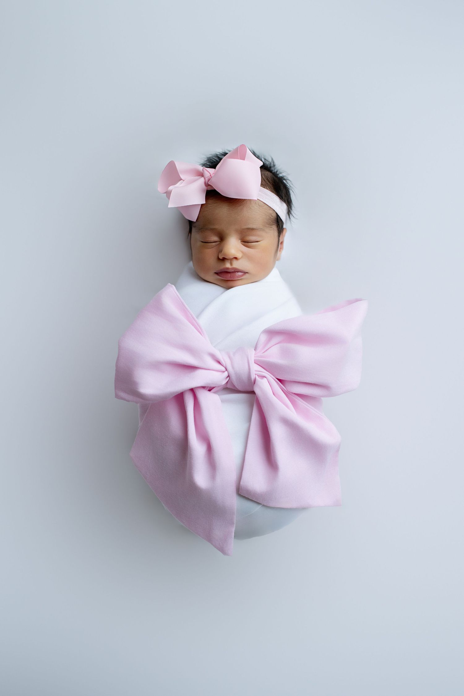 Sleeping newborn baby girl swaddled in pink bow wrap with matching headband on light blue background.