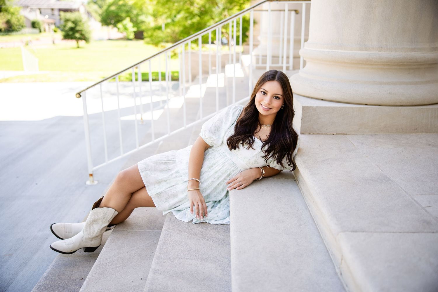 Brunette girl in white lace dress and white boots sitting on stone steps by large column.
