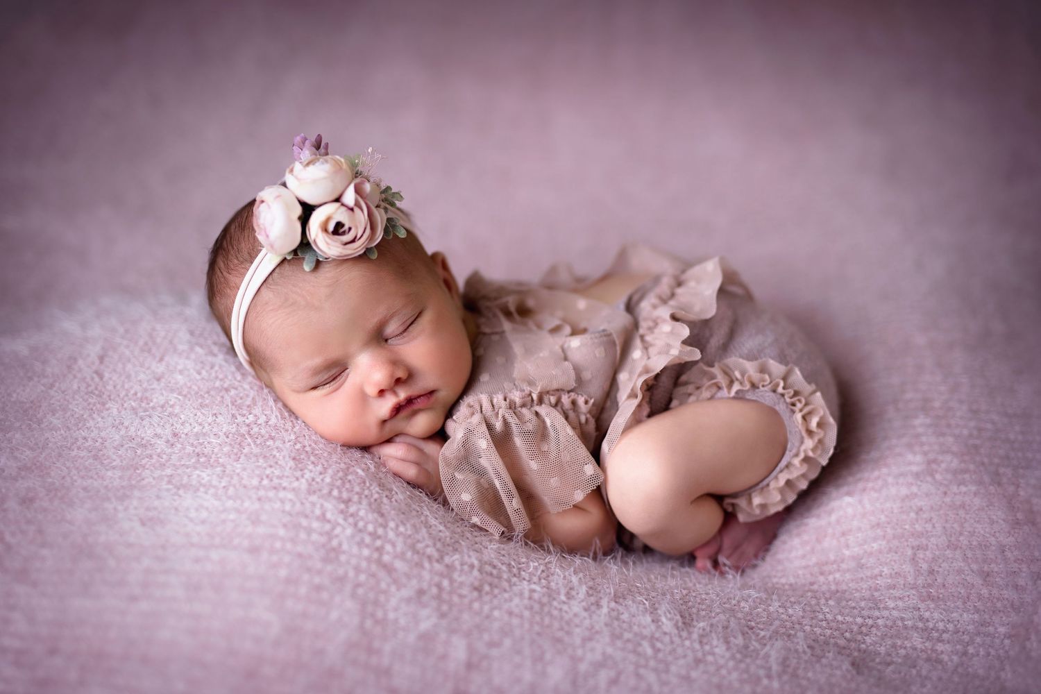 Peaceful newborn baby in lace romper and floral headband sleeps curled up on a soft lavender blanket.