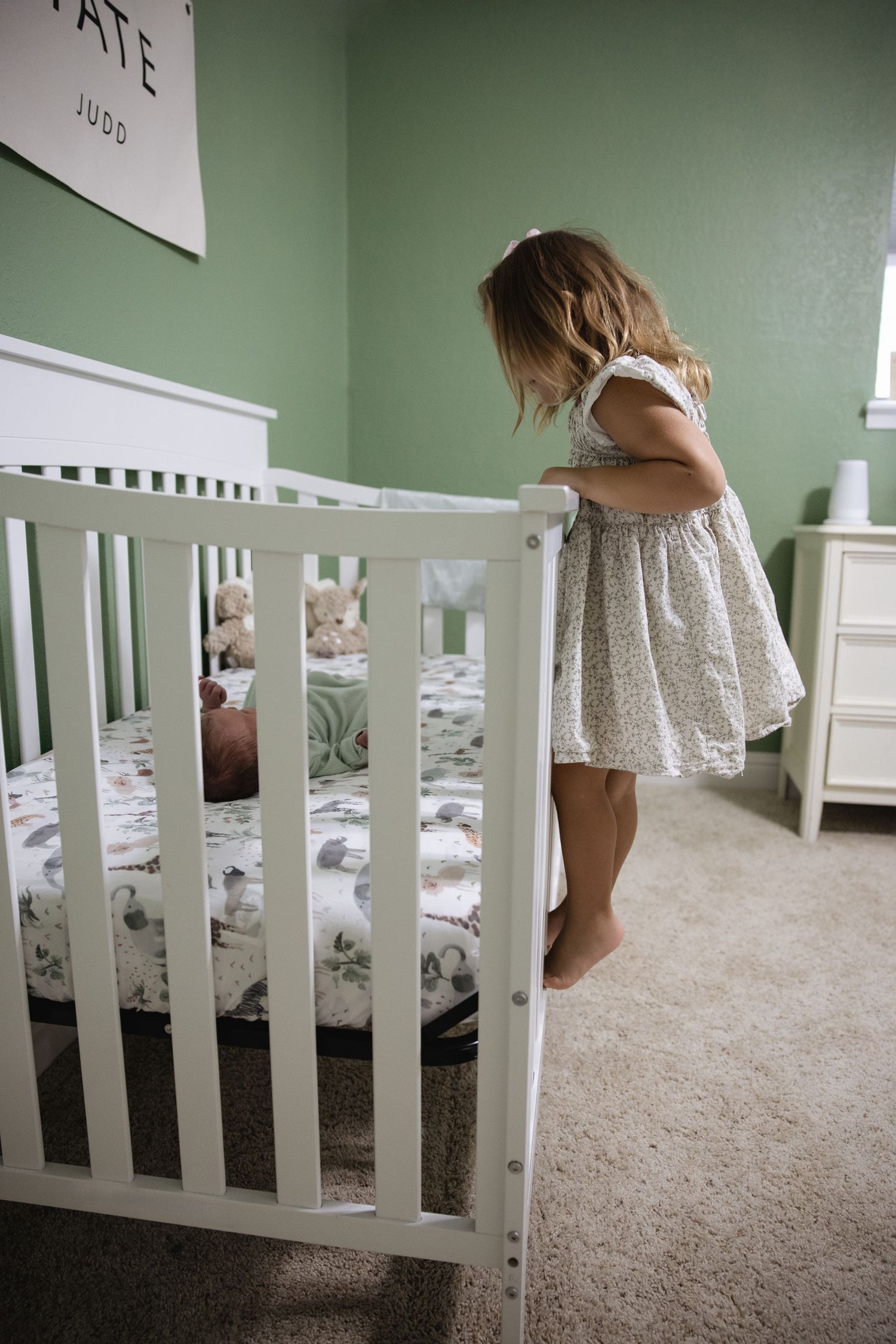 Young girl in floral dress peers into white crib at newborn sibling in green nursery.
