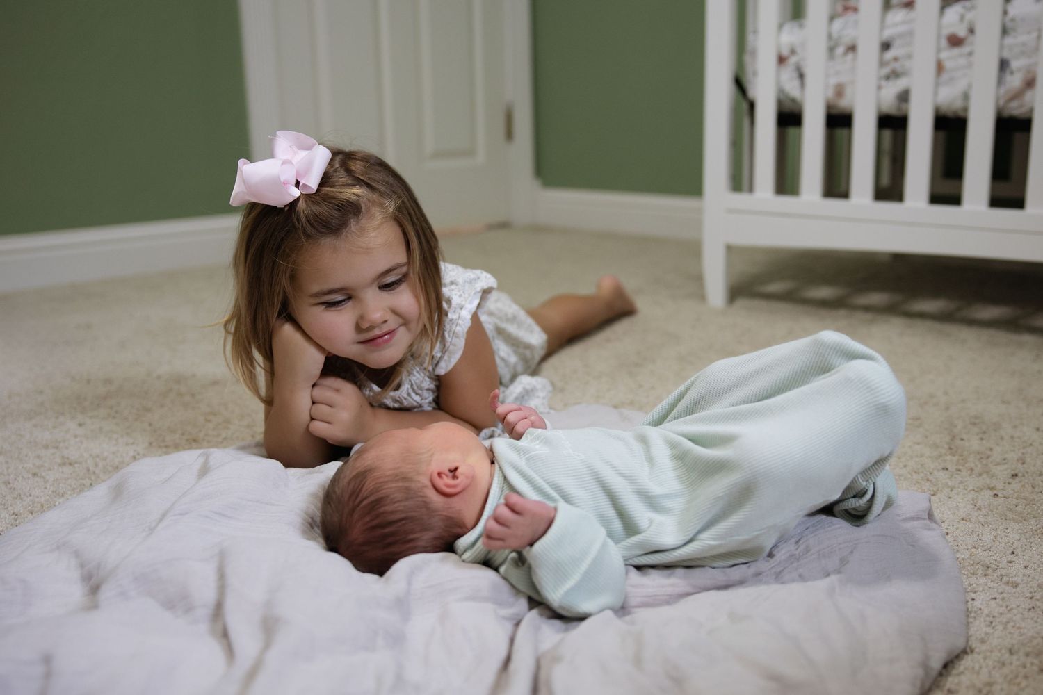 Smiling toddler girl with bow lying next to newborn baby sibling on nursery floor.