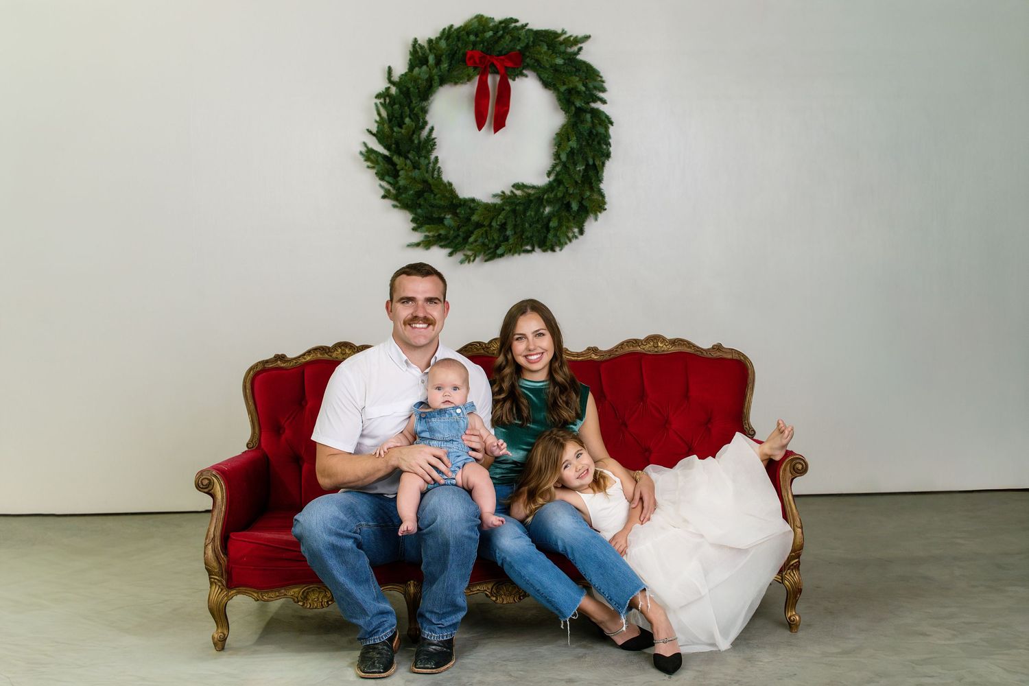 Happy family of four poses on red velvet sofa beneath Christmas wreath in holiday portrait.