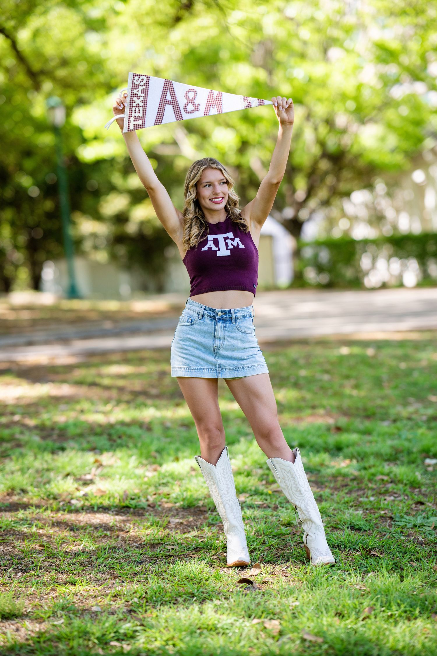 Blonde woman in crop top and denim skirt holding Texas A&M pennant in green park.