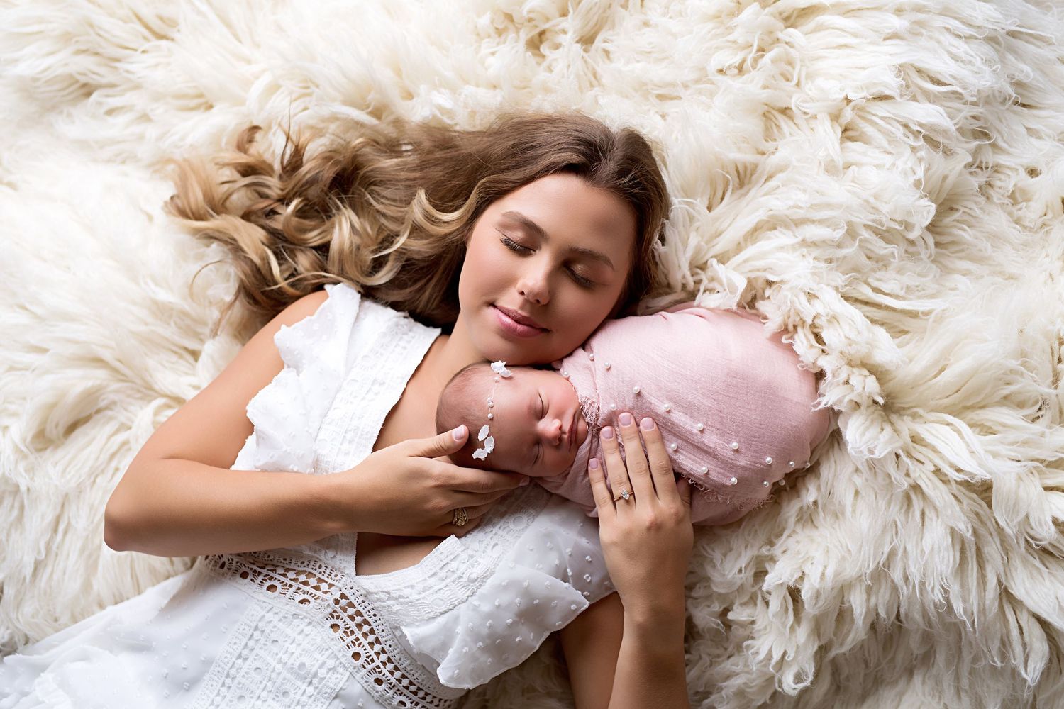 Mother tenderly cuddles sleeping newborn baby on fluffy white fur blanket in professional portrait.