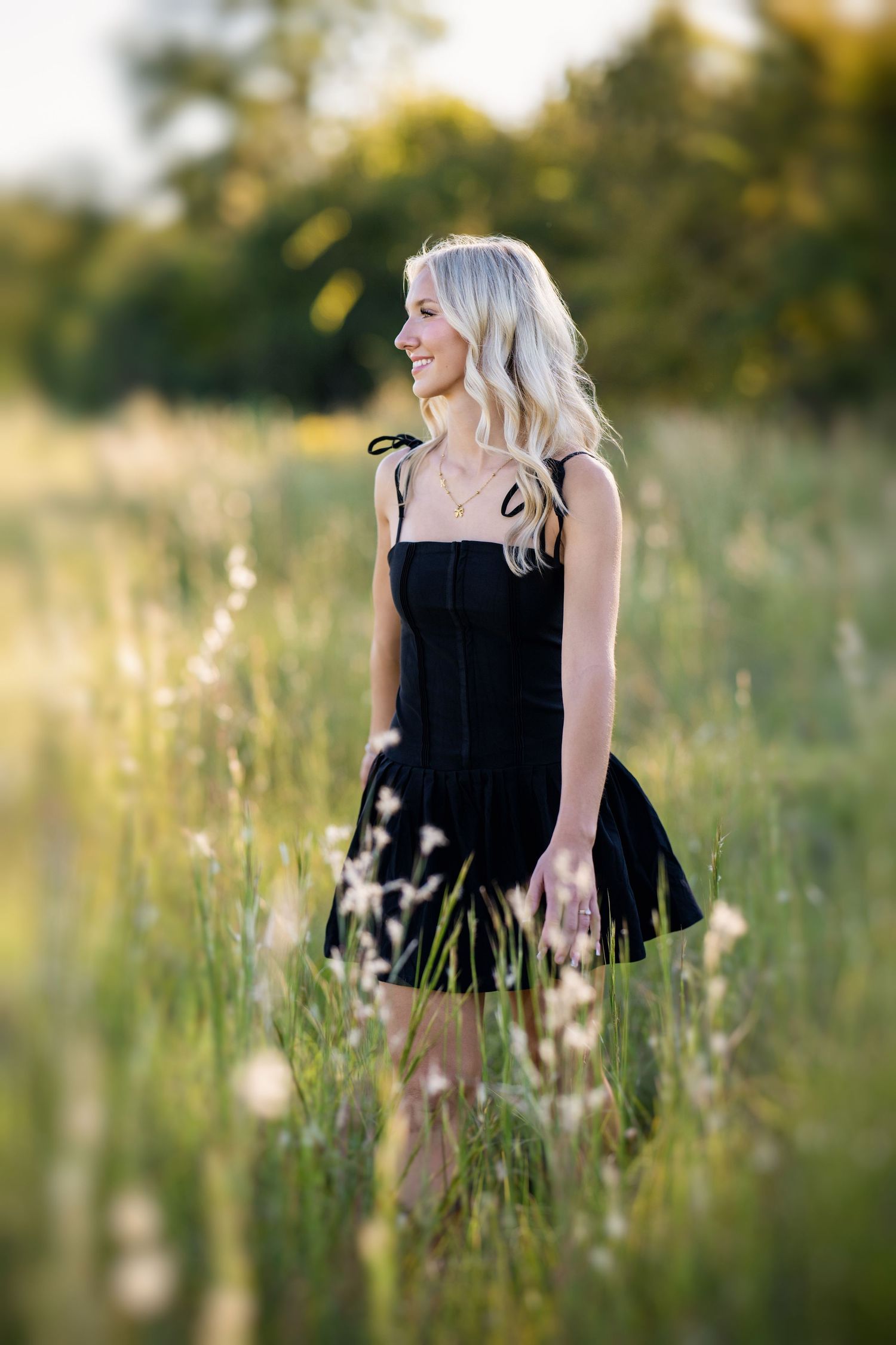 Blonde teen in black sundress smiles softly while standing in a sunlit grassy field.