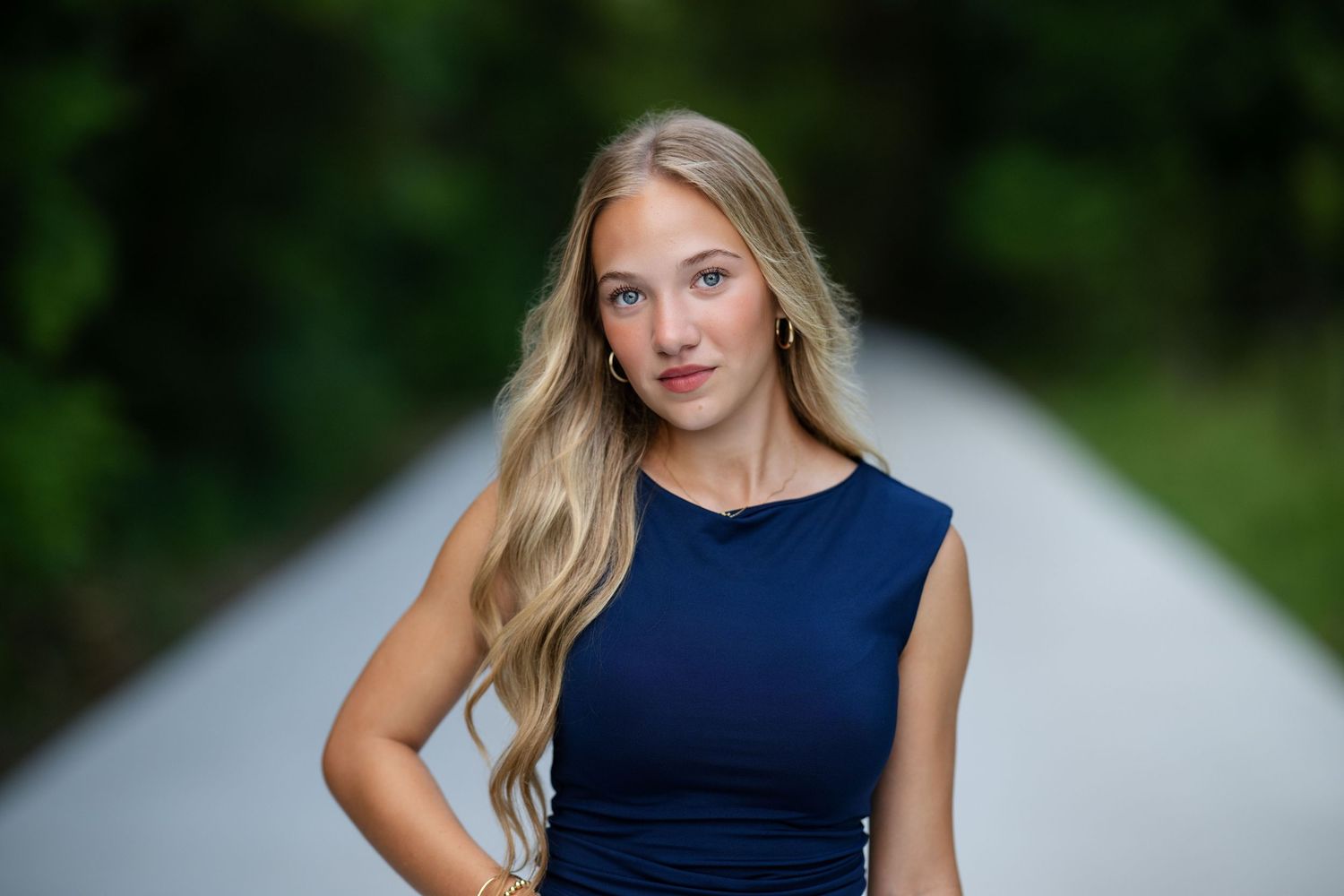 Young woman with long blonde hair poses in navy blue dress on a tree-lined path.