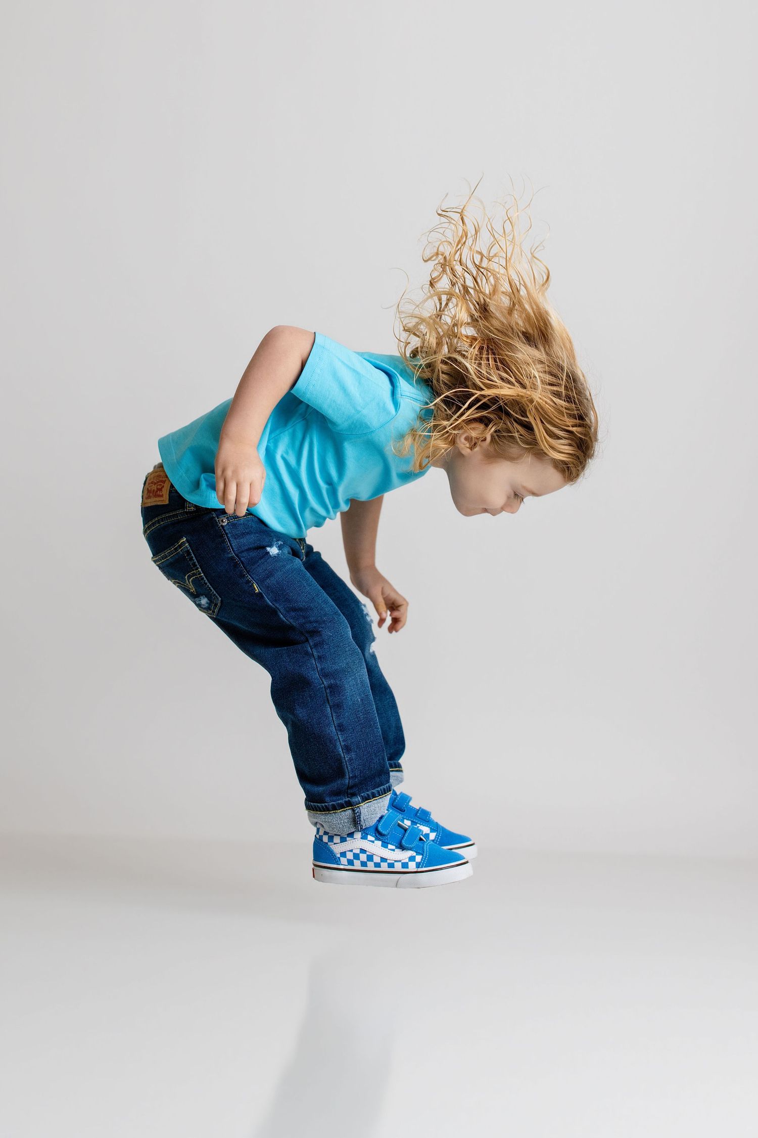 Energetic child with blonde hair jumping in blue sneakers and jeans on white background.