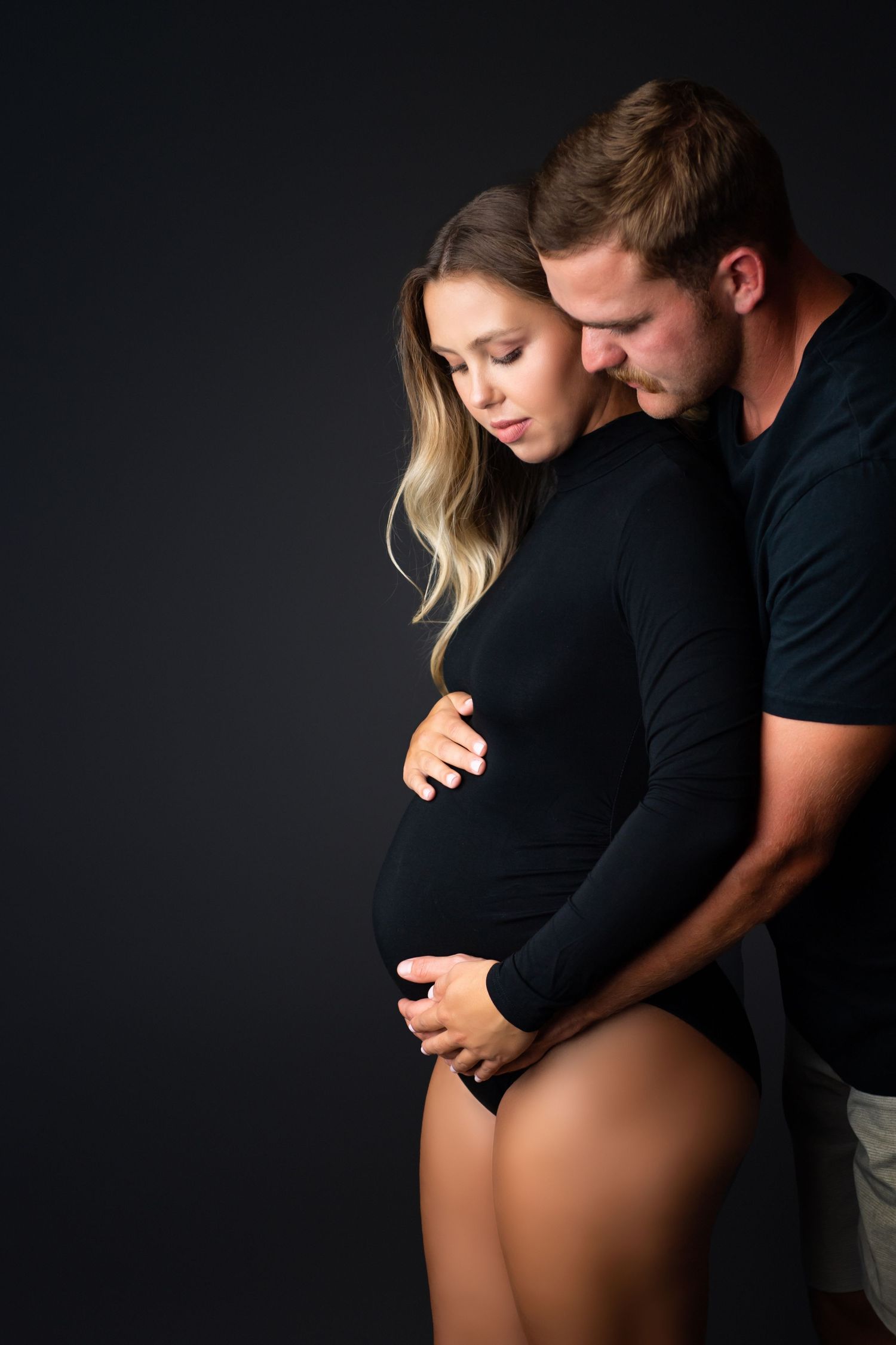 Expecting couple in black outfits tenderly cradling pregnant belly against dark studio background.