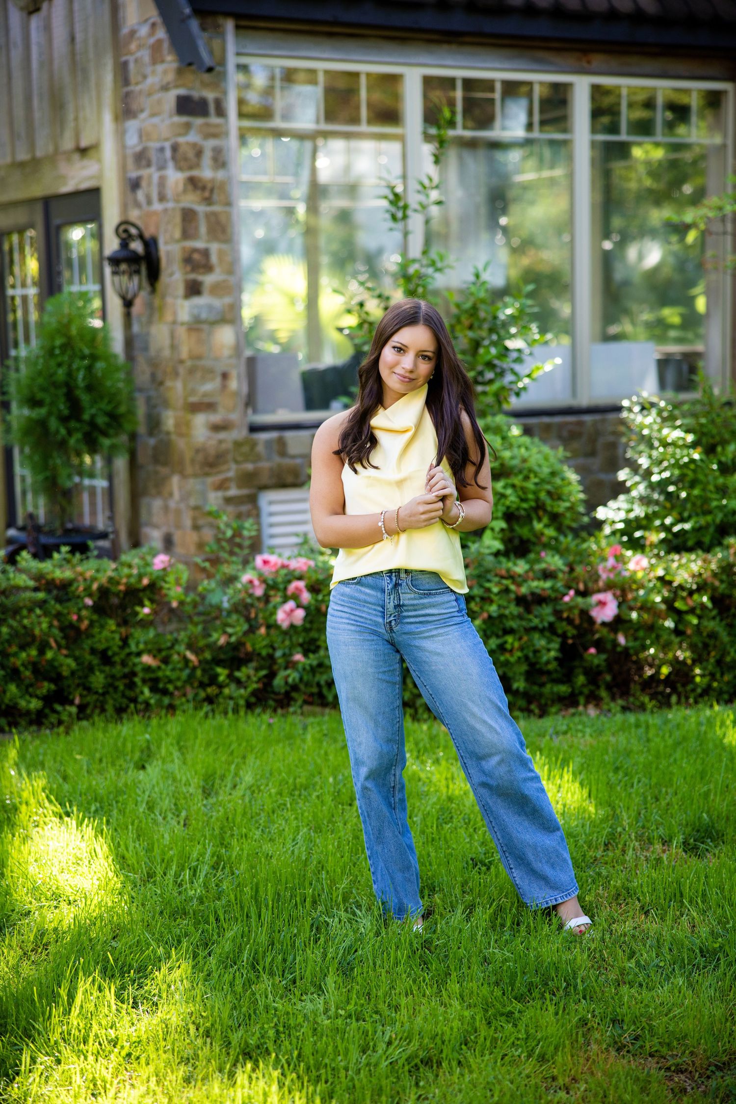 Young woman in yellow halter top and wide-leg jeans poses on green lawn outside brick home.