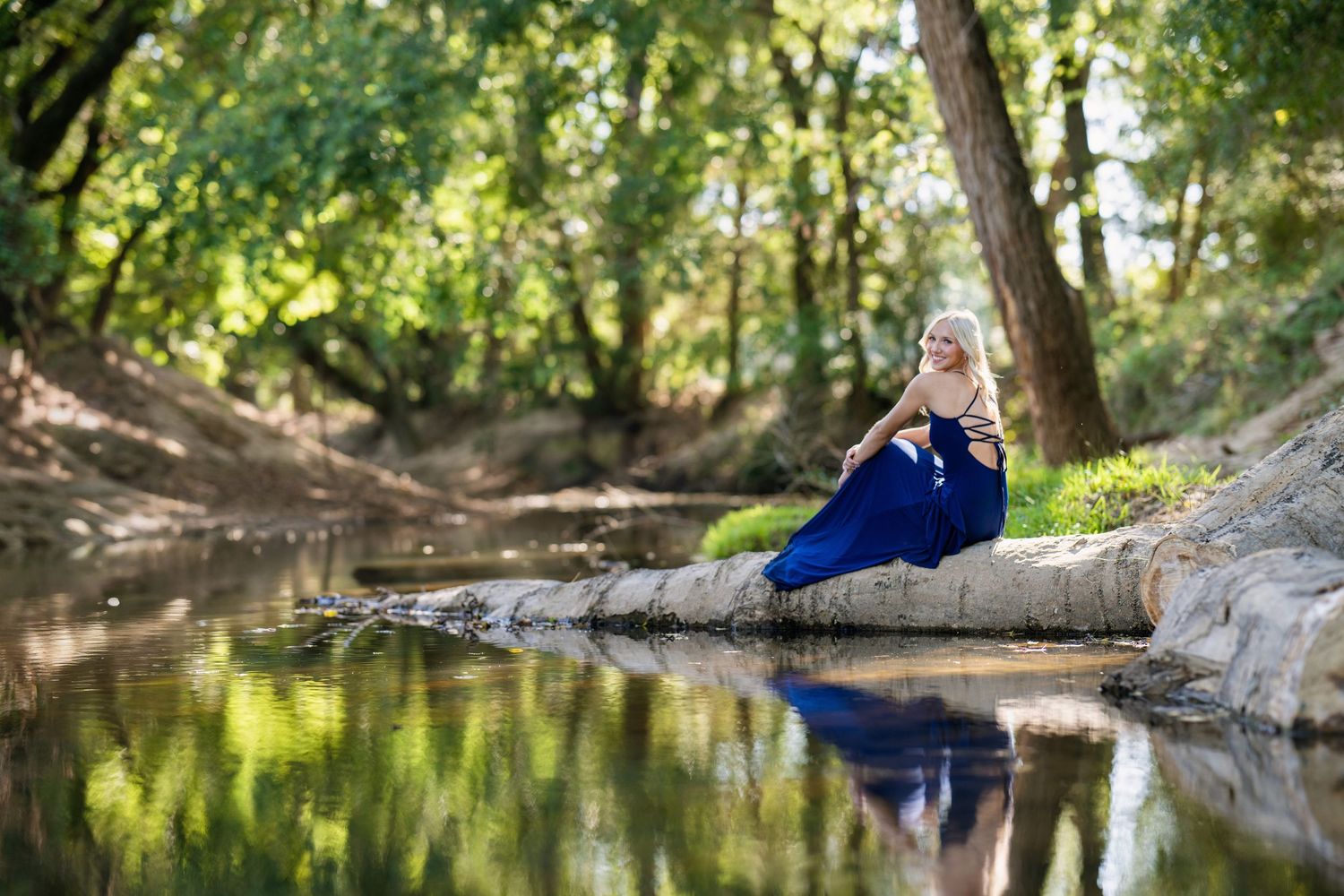 Blonde woman in a navy blue dress relaxing on a log by a tranquil forest creek in sunlight.