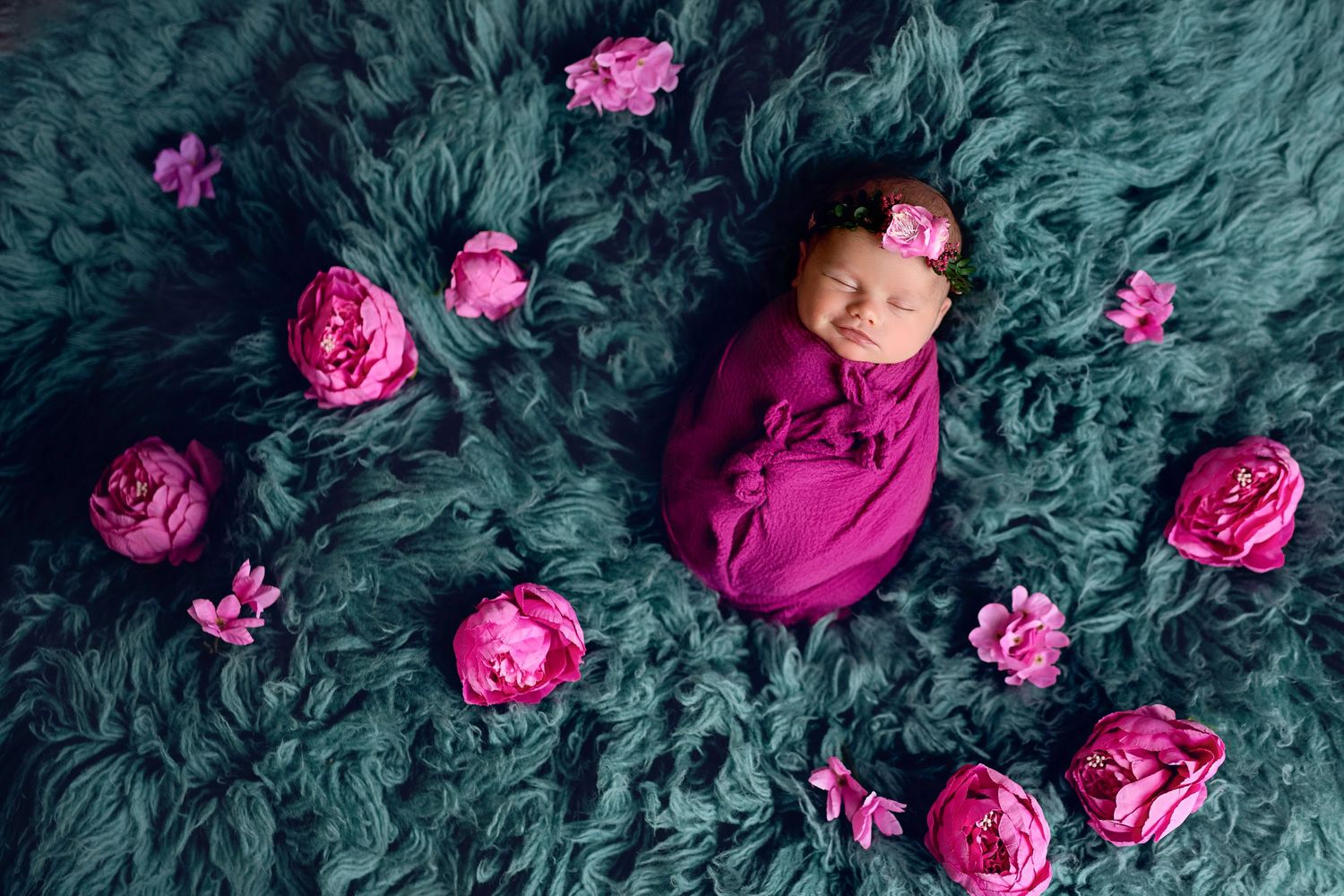 Sleeping newborn wrapped in pink swaddle surrounded by magenta peonies on a teal fur backdrop.