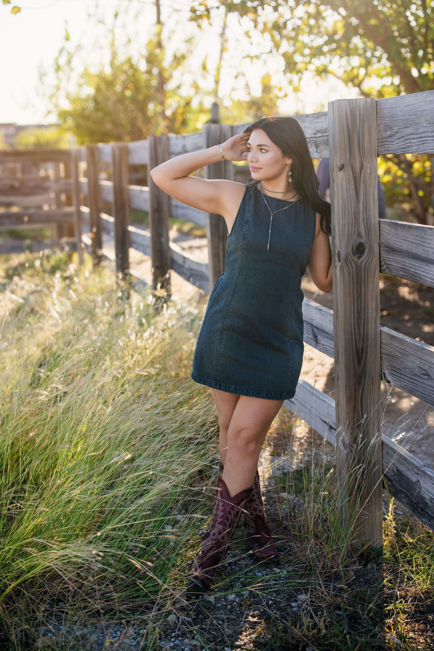 Brunette teen girl in dark sleeveless dress and red cowboy boots leaning on rustic wooden fence.