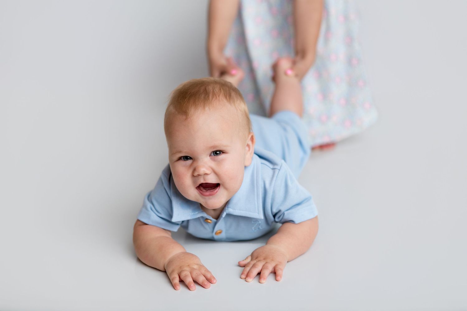 A laughing baby in a blue polo shirt does tummy time on a white studio floor.