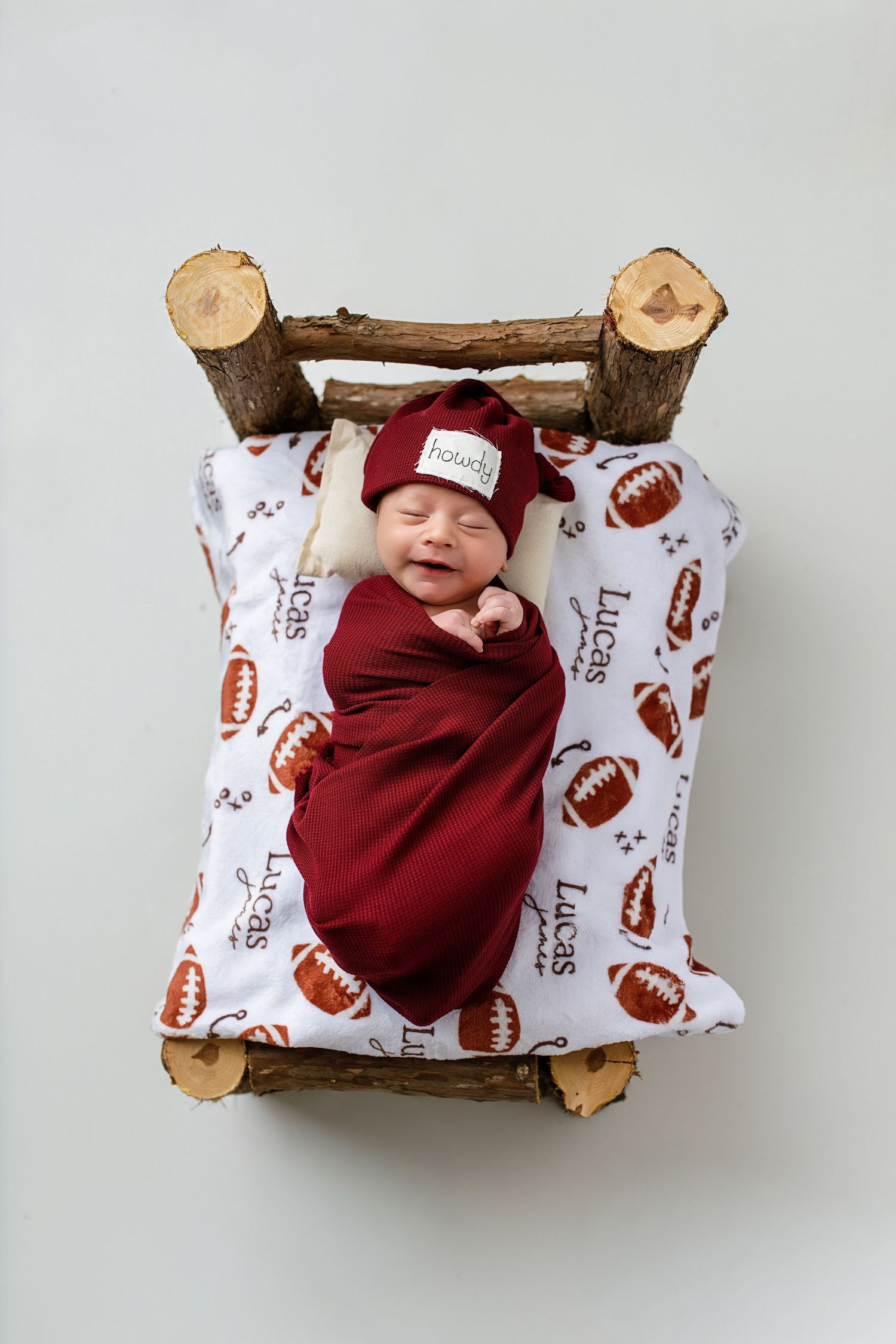 Smiling newborn named Lucas swaddled in burgundy wrap lies in a rustic log bed with football-print blanket.