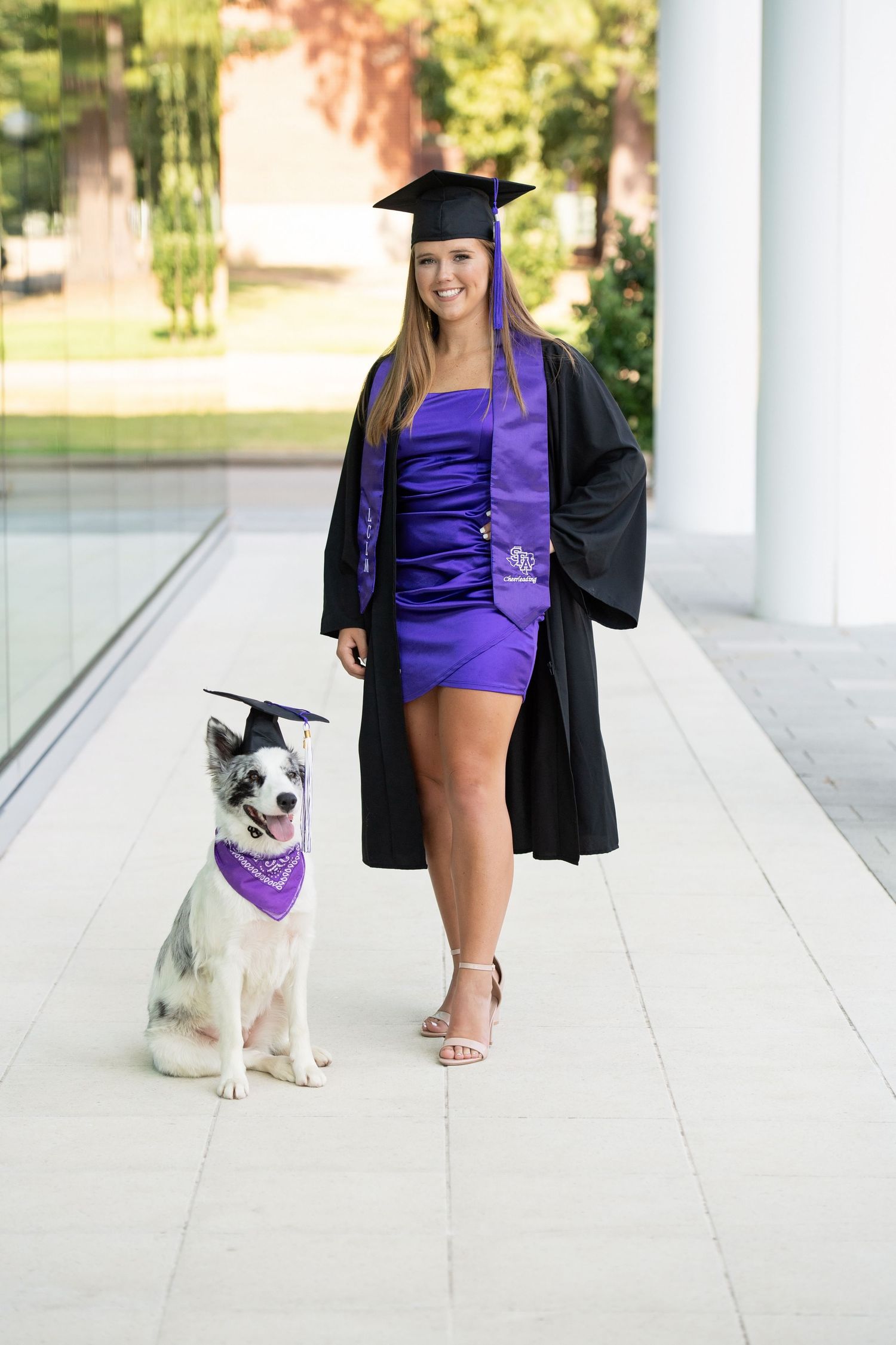 Smiling graduate in purple dress and black cap and gown posing with a dog in matching graduation cap.