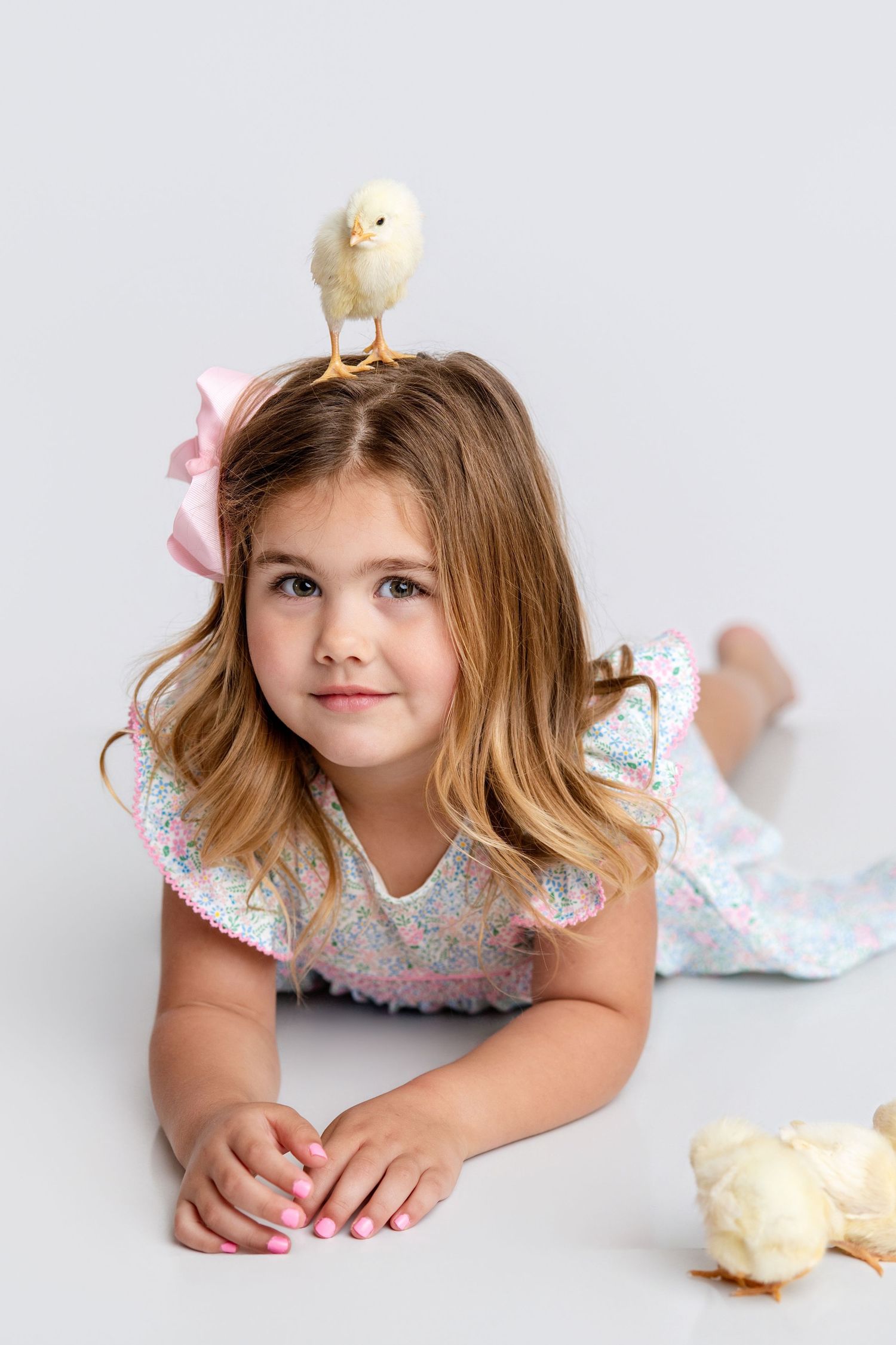 A young girl in a floral dress lies on the floor with a baby chick perched on her head.