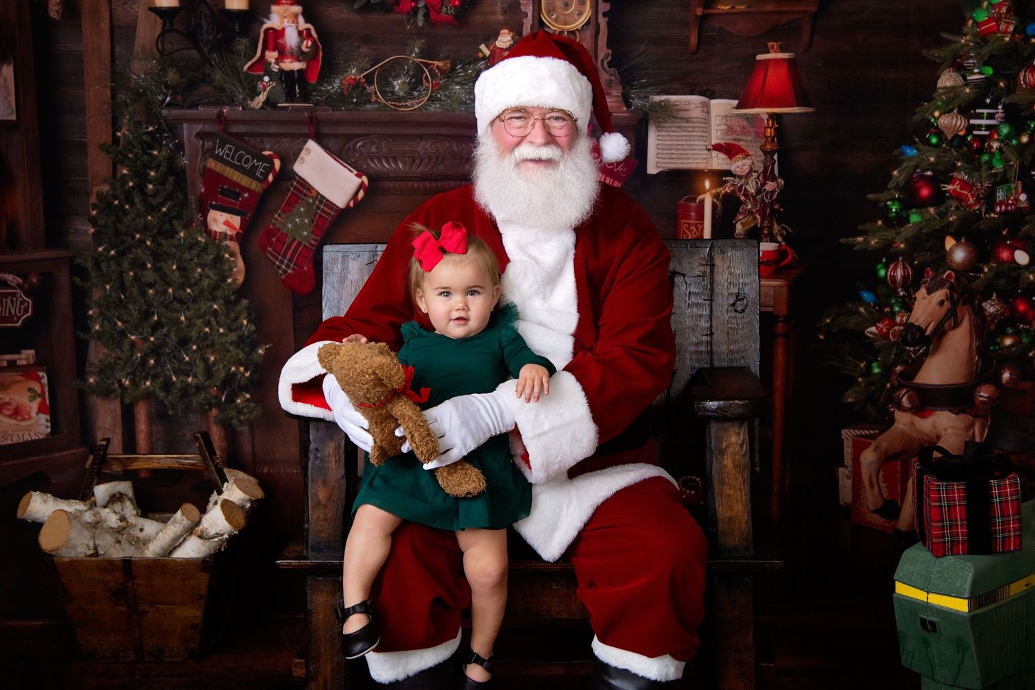 Smiling baby girl in green dress sits on Santa Claus lap holding a teddy bear in a festive Christmas setting.