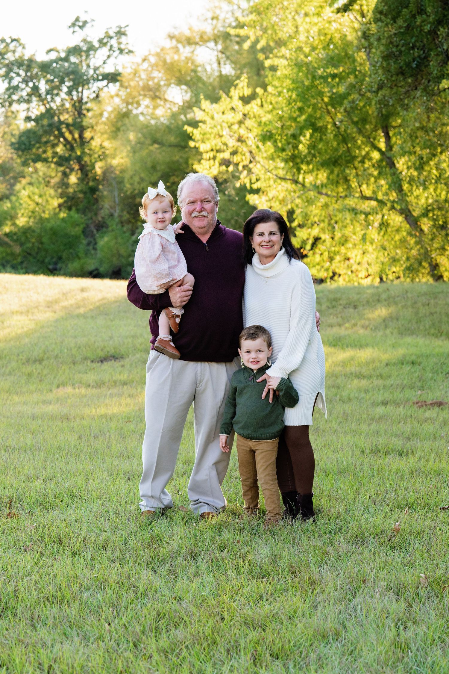 Grandparents smiling outdoors in a green field, holding a baby girl and standing with a young boy.