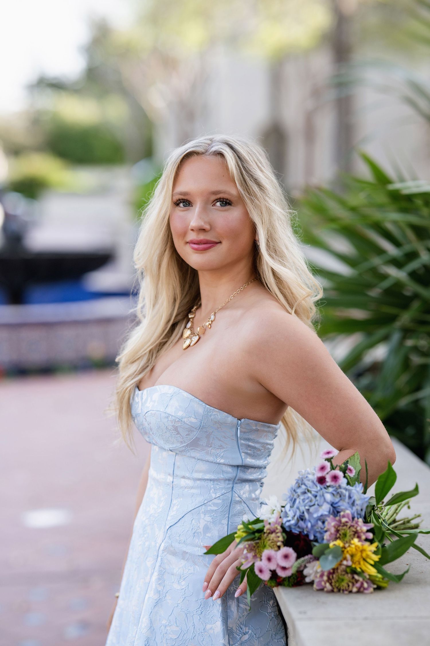 Blonde teen in light blue strapless gown holds flowers for elegant senior portrait outdoors.
