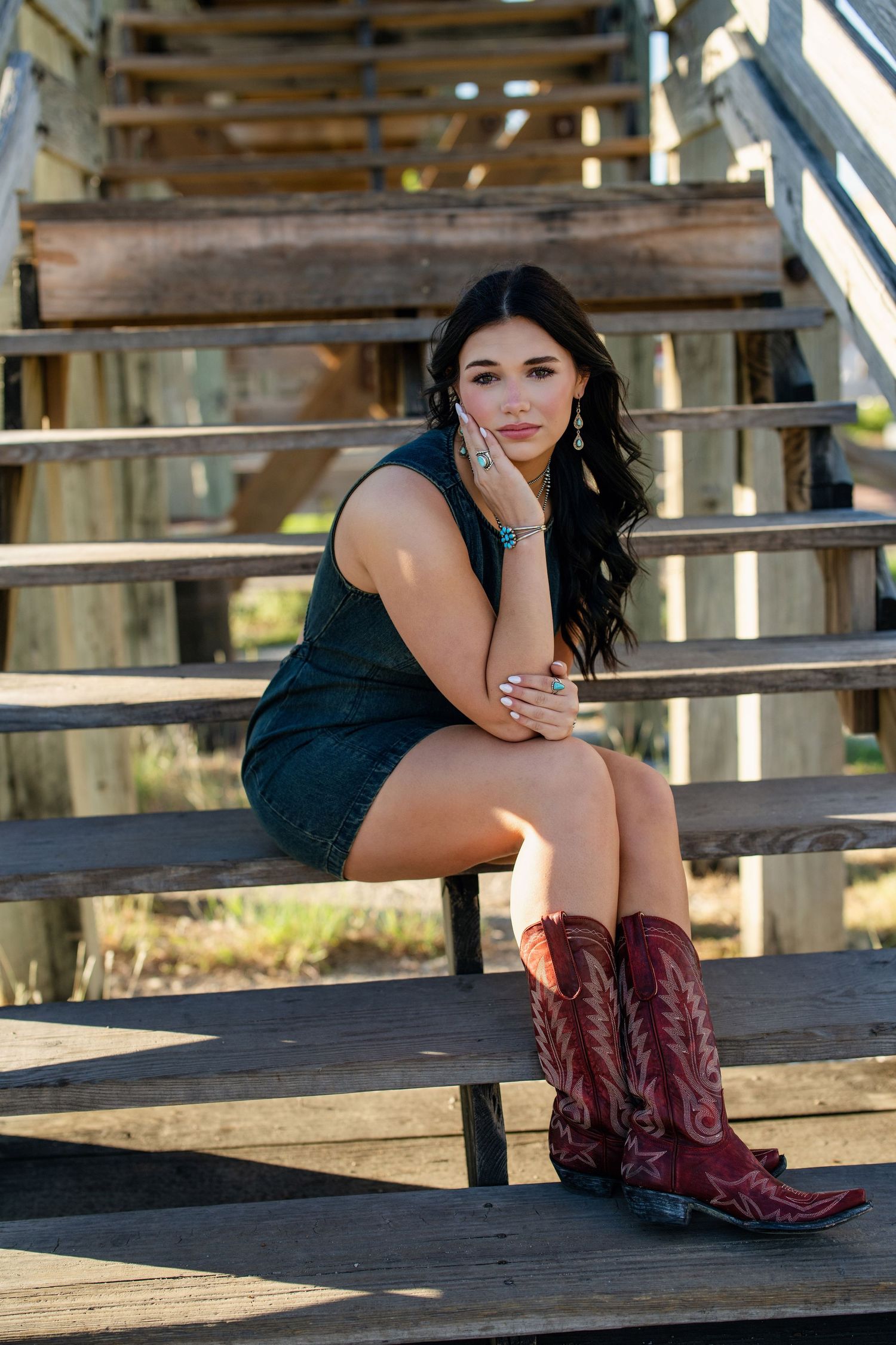 Dark-haired woman in teal mini dress and red cowboy boots poses on wooden bleachers.